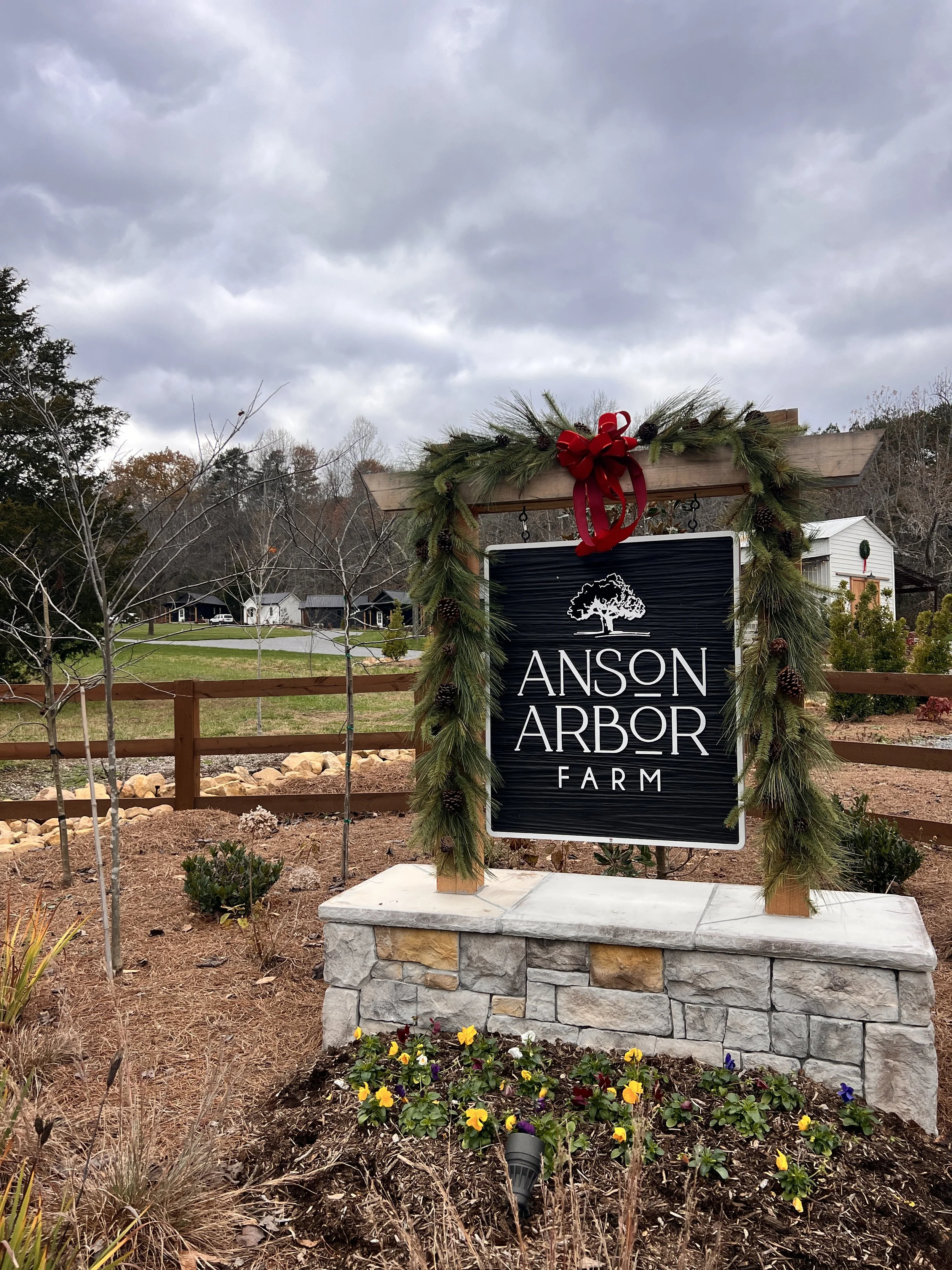 Entrance sign for Anson Arbor Farm surrounded by festive pine garlands and a red bow, set against a cloudy sky backdrop, with landscaped grounds and trees in the background.