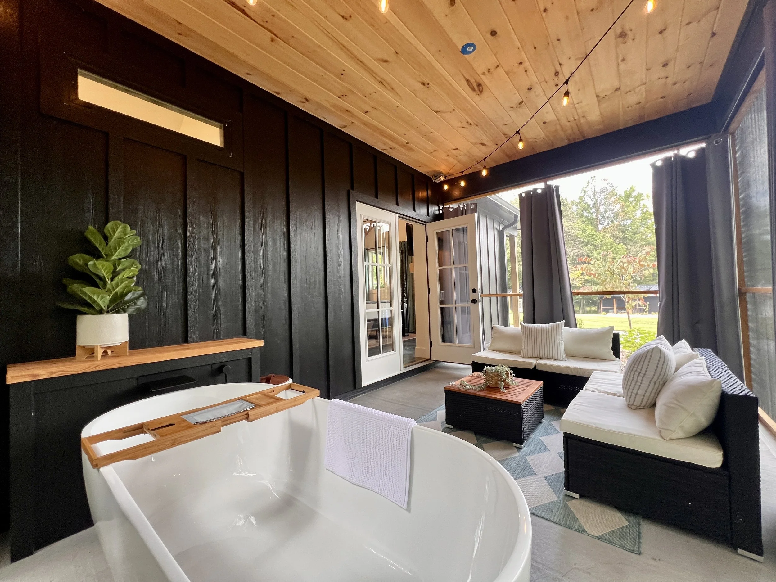 Cozy outdoor patio with black walls and wood ceiling, featuring a freestanding white bathtub with a wooden tray and towel, a potted plant on a wooden shelf, a wicker sofa set with white cushions, a coffee table, string lights, and a view of a garden.