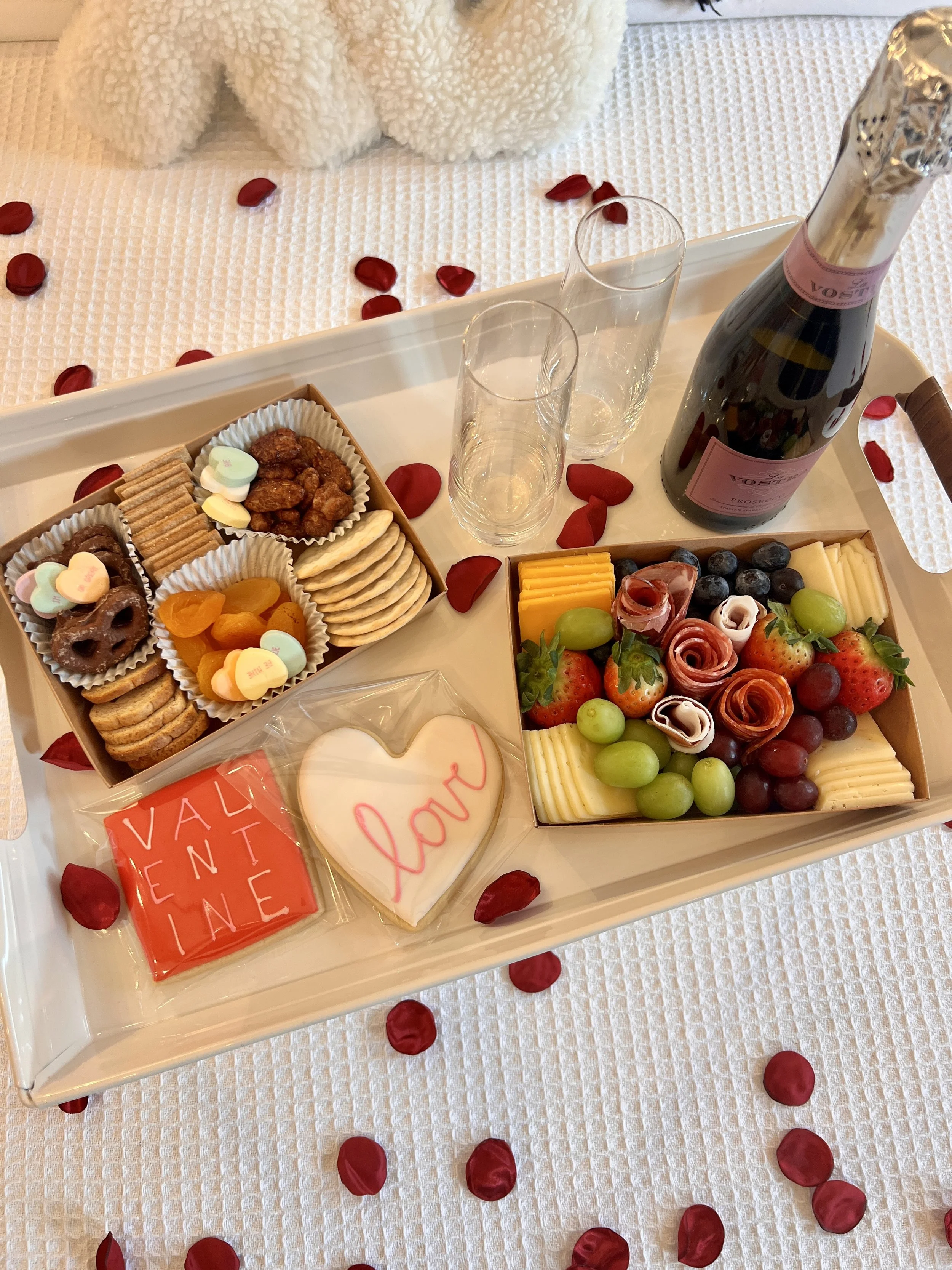 A tray with Valentine's Day-themed items including two compartments with crackers, cheese, nuts, dried fruits, strawberries, grapes, ham, and heart-shaped chocolates. There are two decorated cookies, one square with 'Valentine' and one heart-shaped w