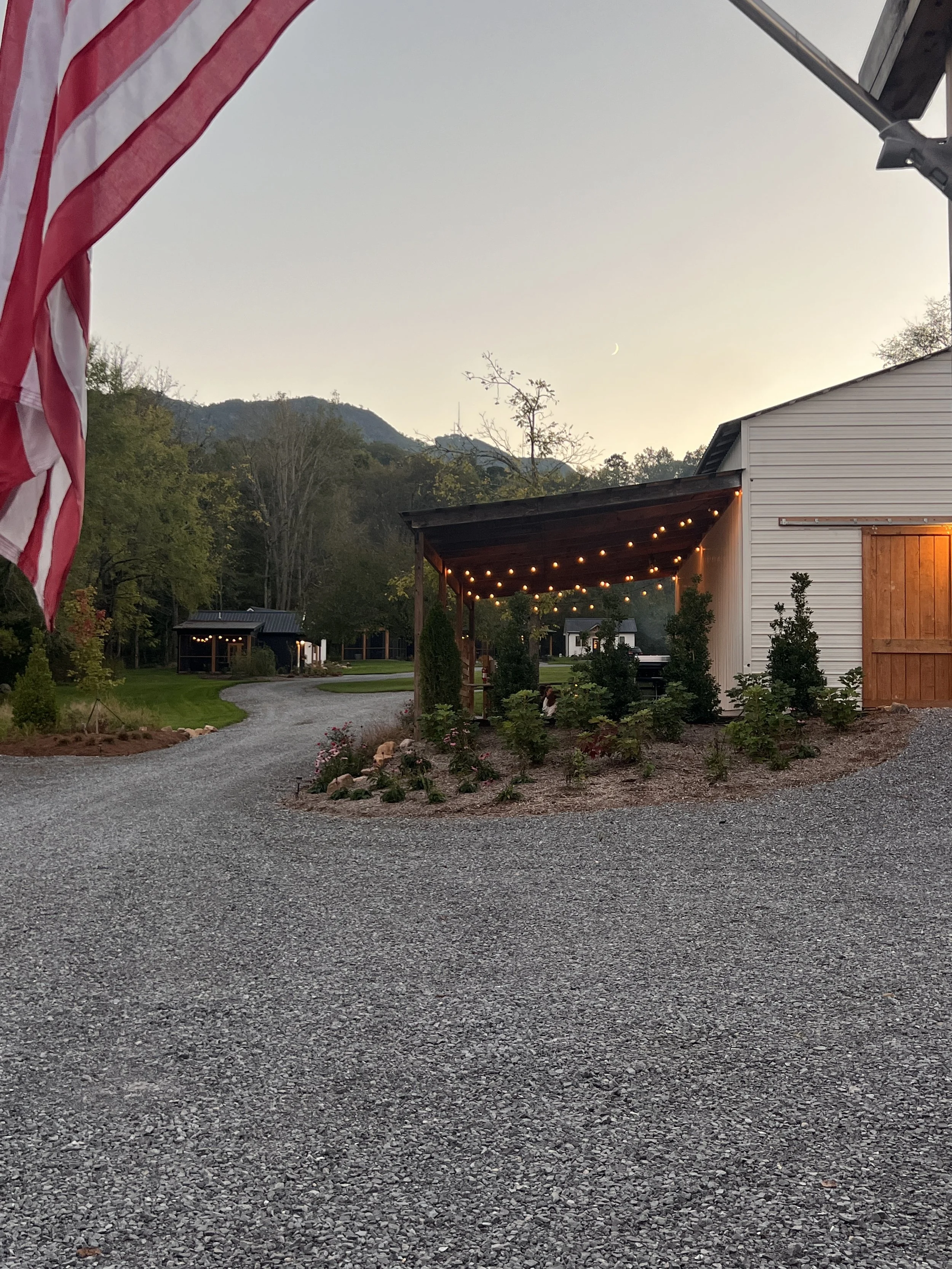 Gravel driveway leading to a building with string lights on a porch, surrounded by trees and mountains in the background; an American flag is partially visible in the foreground.
