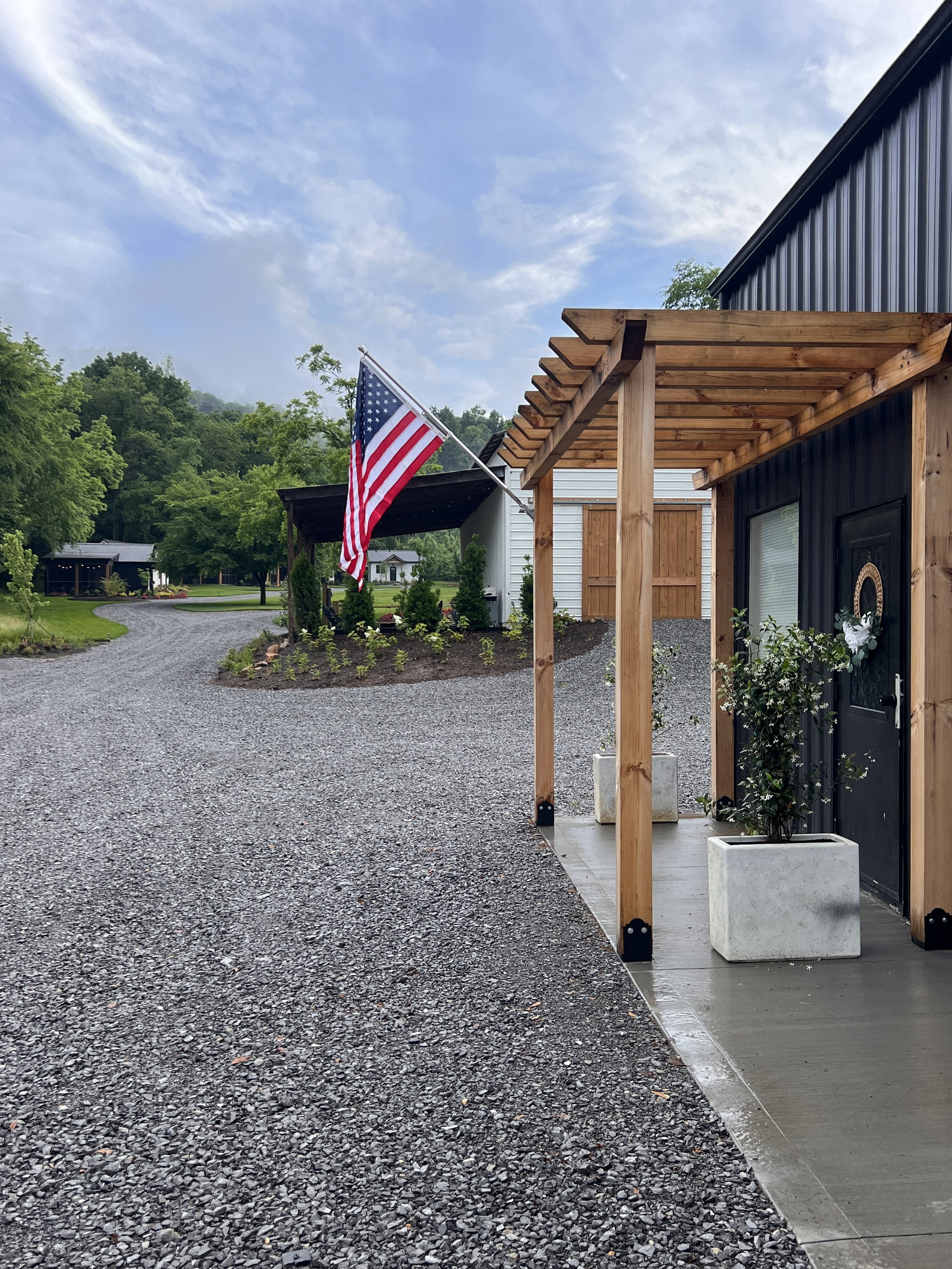 A building with a wooden pergola and an American flag on display. The area is surrounded by gravel, greenery, and trees, with a driveway leading to another building in the background under a cloudy sky.