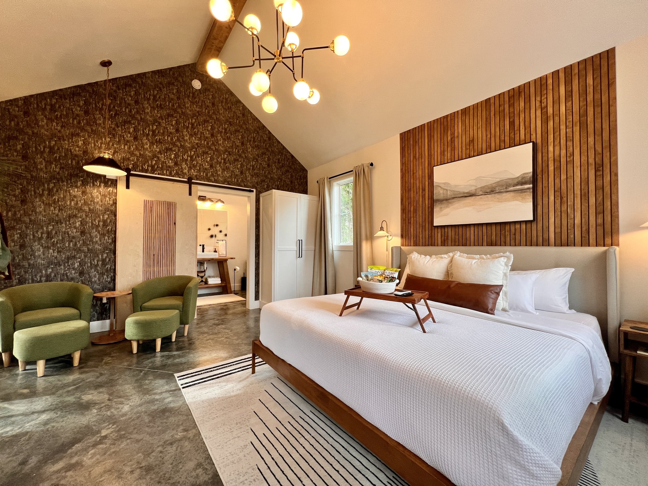 Modern bedroom with a king-sized bed, white bedding, and decorative pillows. Wooden accent wall with landscape artwork above bed. Two green armchairs and a small wooden table near the bed. Vaulted ceiling with pendant lighting and a sliding barn door