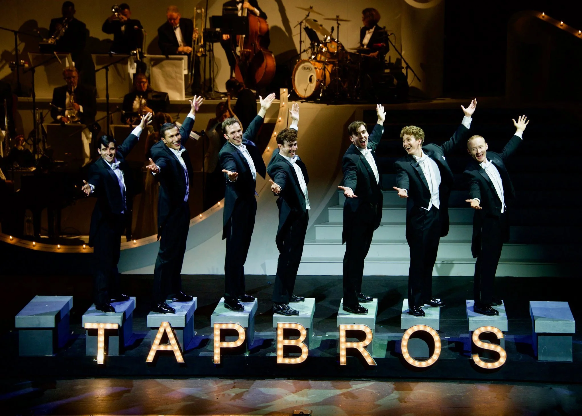 A group of six men in tuxedos performing a choreographed dance on stage, with a lit-up "TAP BROS" sign in front and an orchestra in the background.