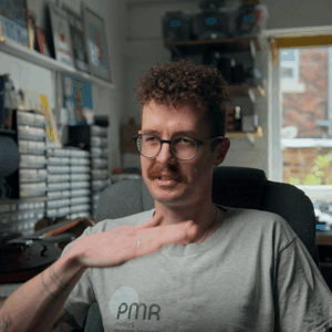 A man with curly hair and glasses sitting in a home office, gesturing with his right hand, with shelves and windows in the background.