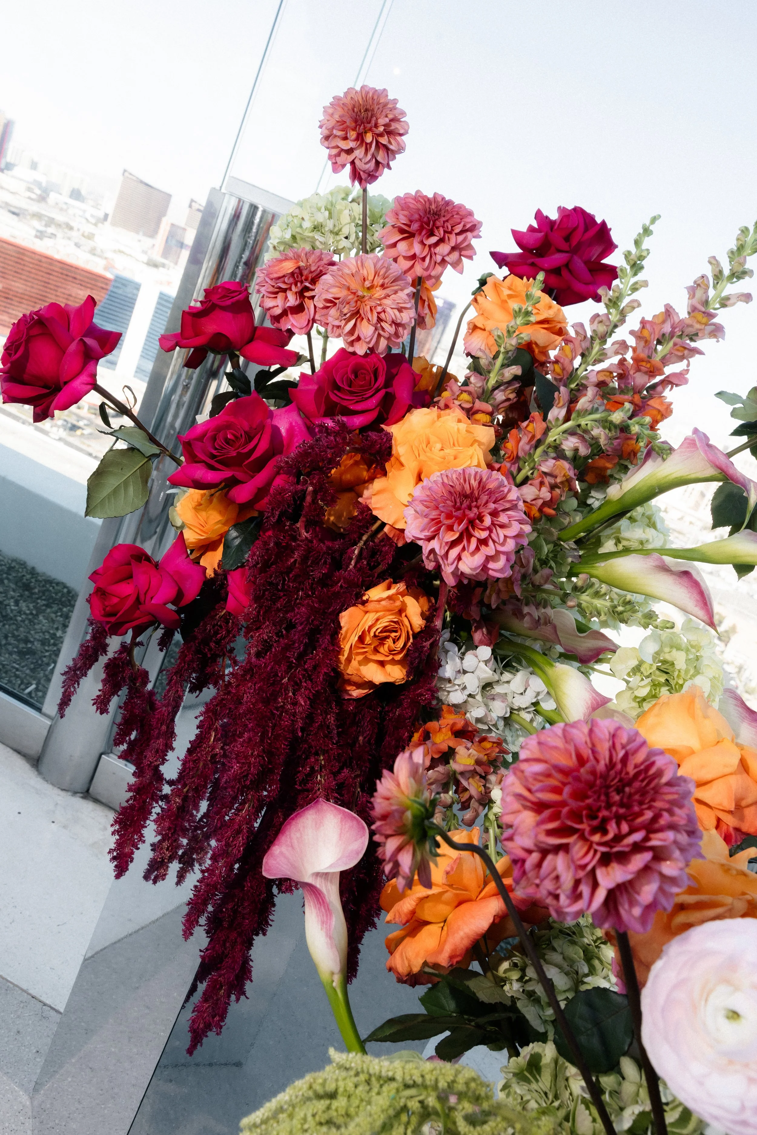 Tall jewel-toned arrangement of burgundy roses, dahlias, orange roses and trailing amaranthus against the Las Vegas skyline, wedding florals by Thistle House