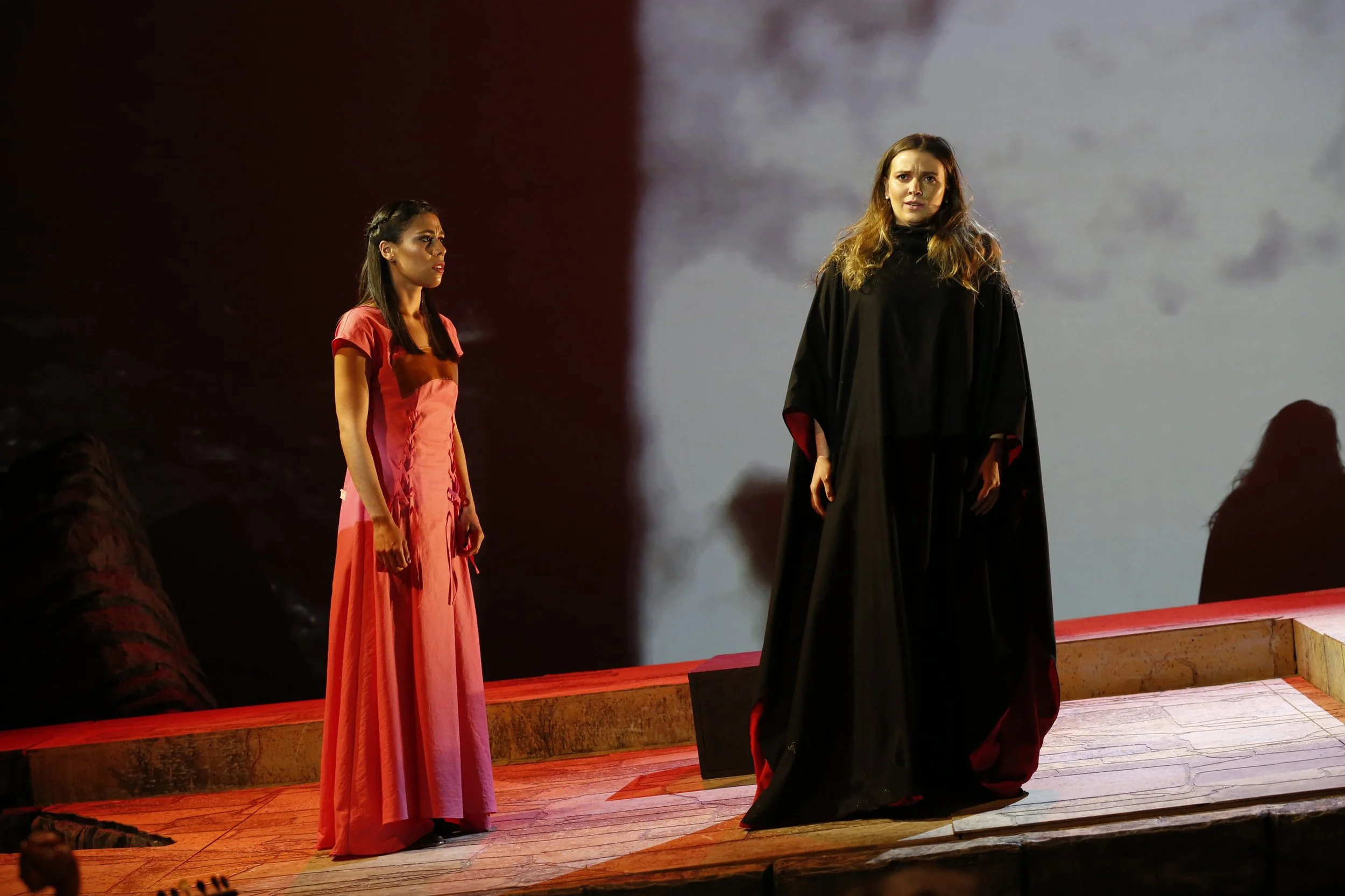 Two women standing on a stage, one in a red dress looking at the other in a black robe, with a cloudy sky projected in the background.