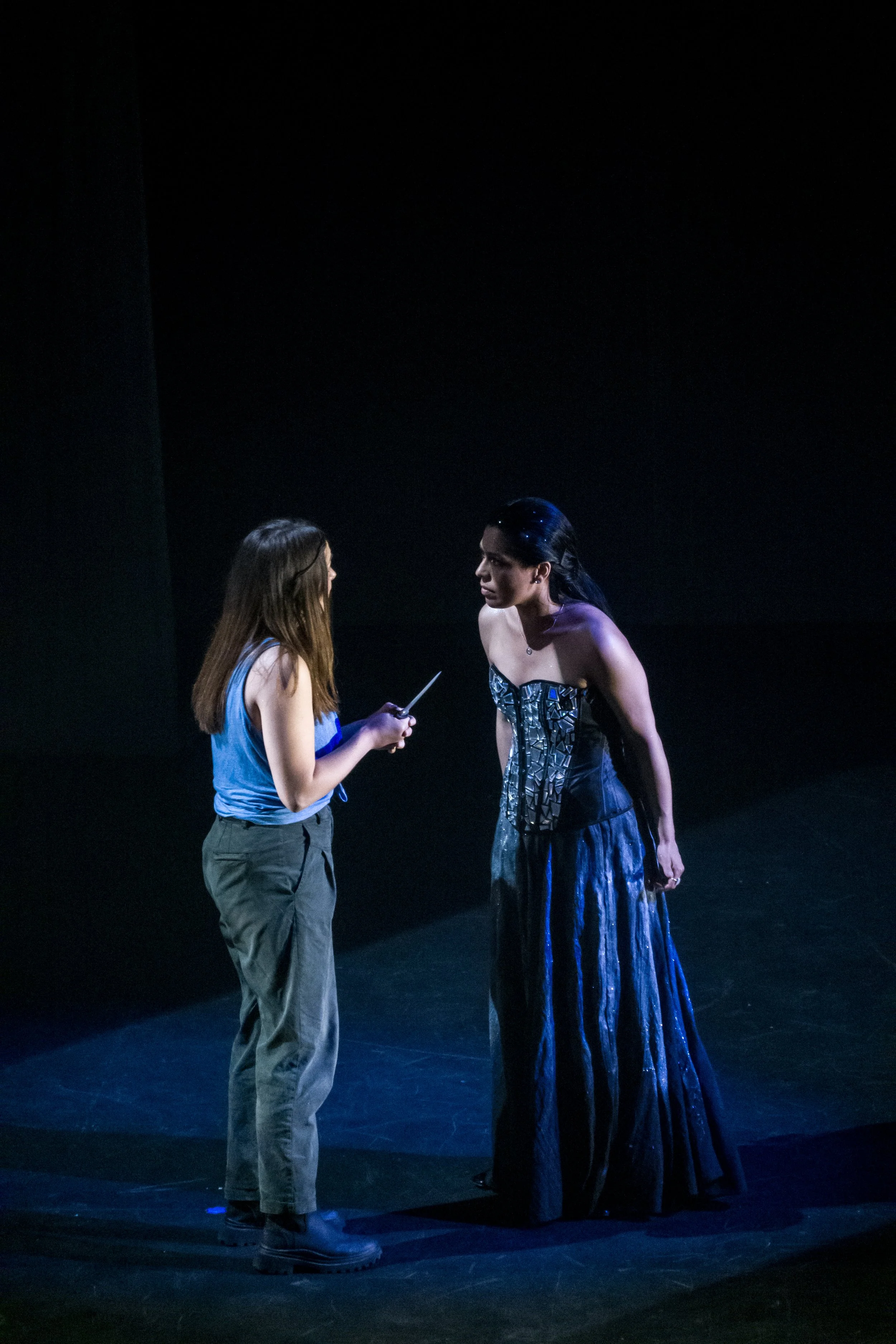 Two women are engaged in a serious conversation on a dark stage. One is wearing a casual outfit of a blue top and tan cargo pants, holding a small object. The other is dressed in an elaborate, metallic, strapless gown with a long, flowing skirt, and 