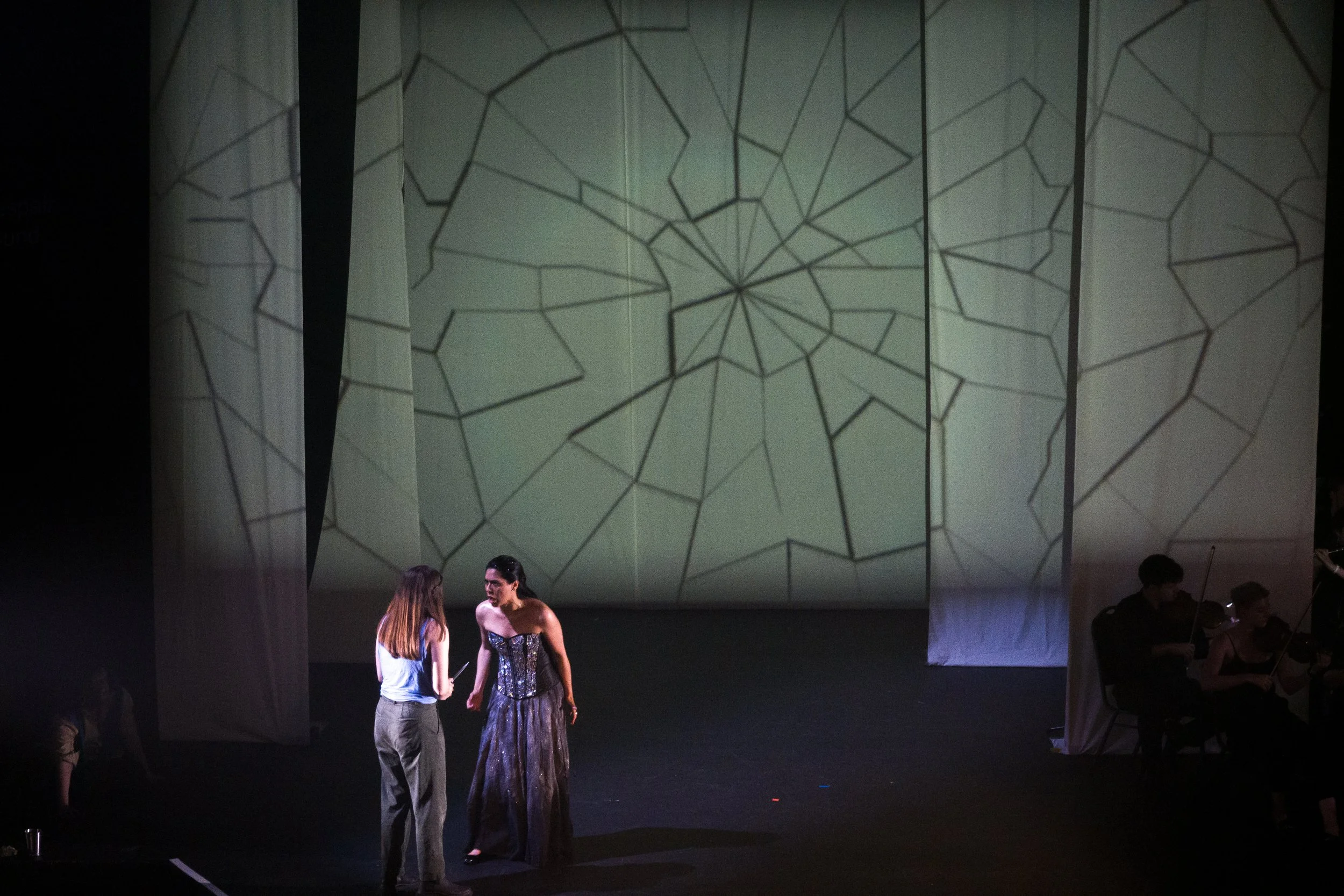 Two women are standing and talking to each other on a dark stage. One woman is in a shiny dress, and the other woman is wearing casual clothing. In the background, there is a large screen with a geometric pattern projection.
