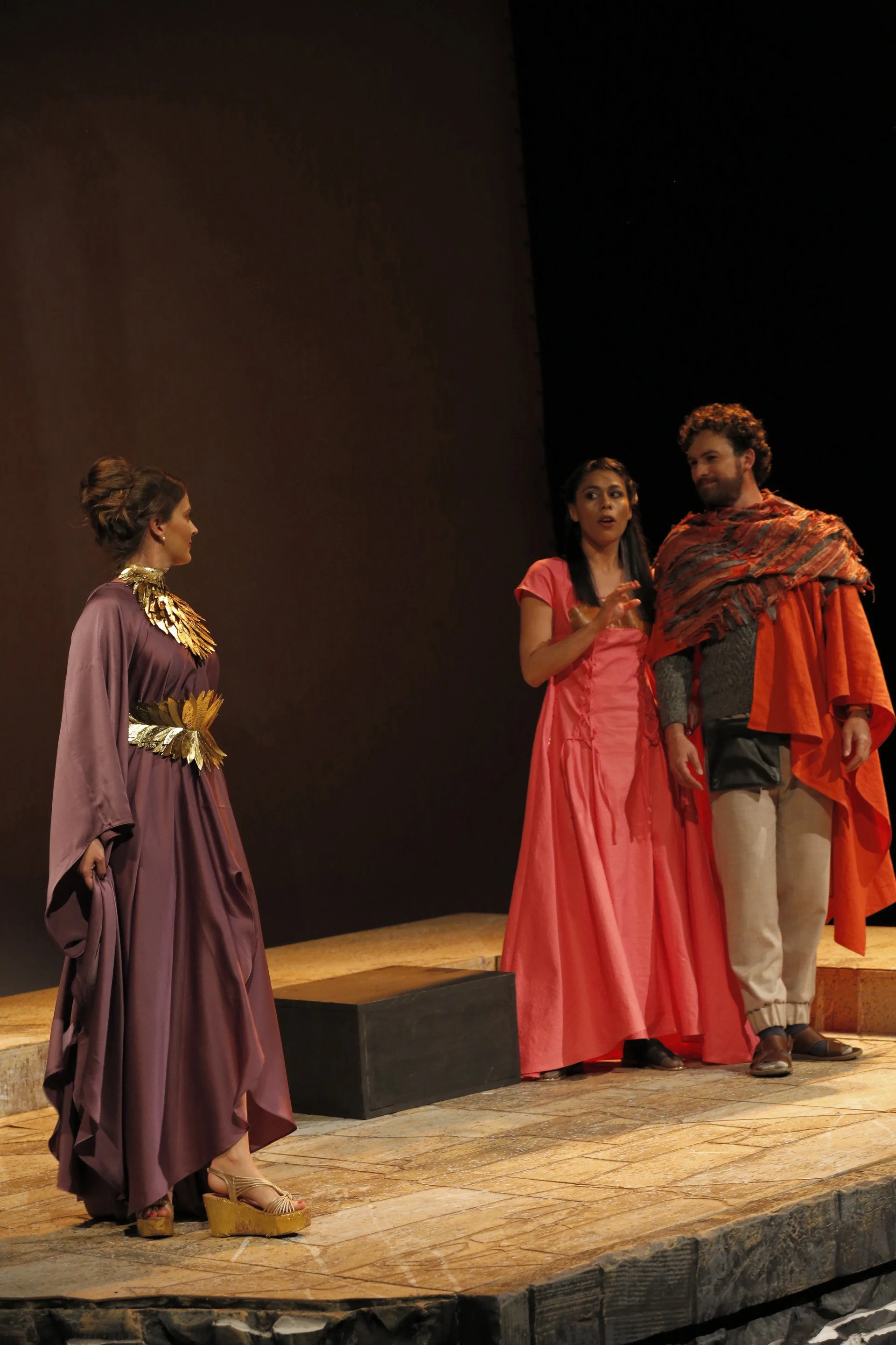 Three actors on stage in a theatrical performance, with one woman in a purple gown with gold accents and platform sandals, speaking to a man and woman dressed in colorful, ancient-inspired costumes, under stage lighting.