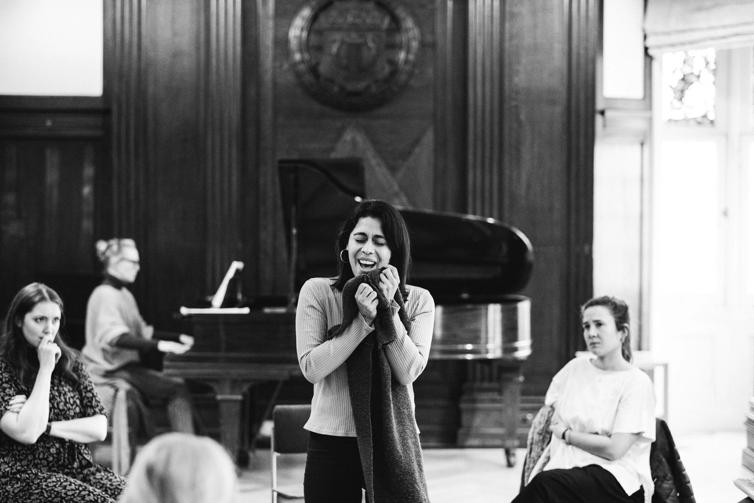 A woman with short dark hair singing or speaking passionately with her eyes closed at a gathering in a room with wood-paneled walls and a grand piano in the background. Other women are sitting or standing around, some listening intently.