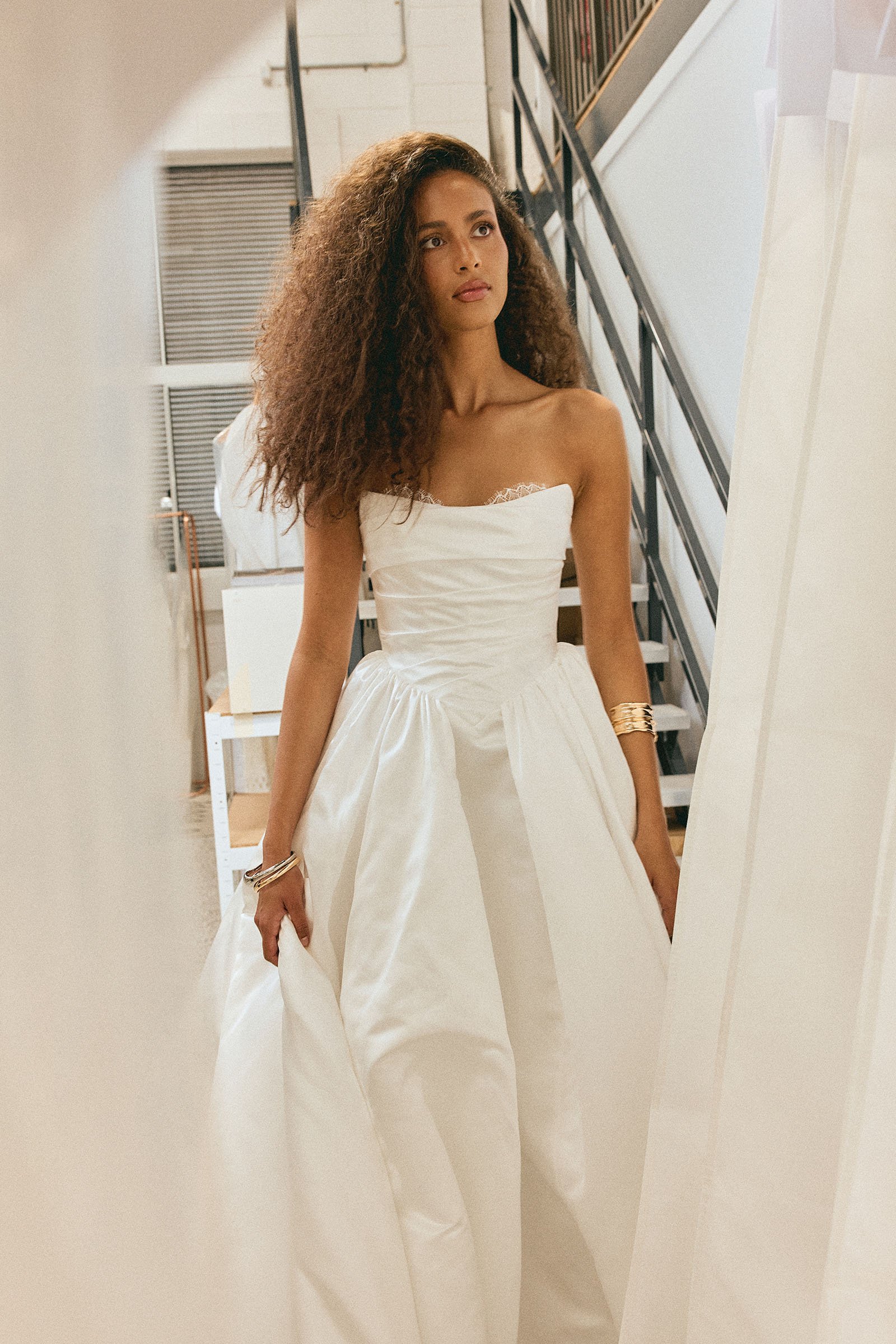 A woman with long curly hair wearing a strapless white wedding dress, standing in a room with metal stair railings and a staircase in the background.