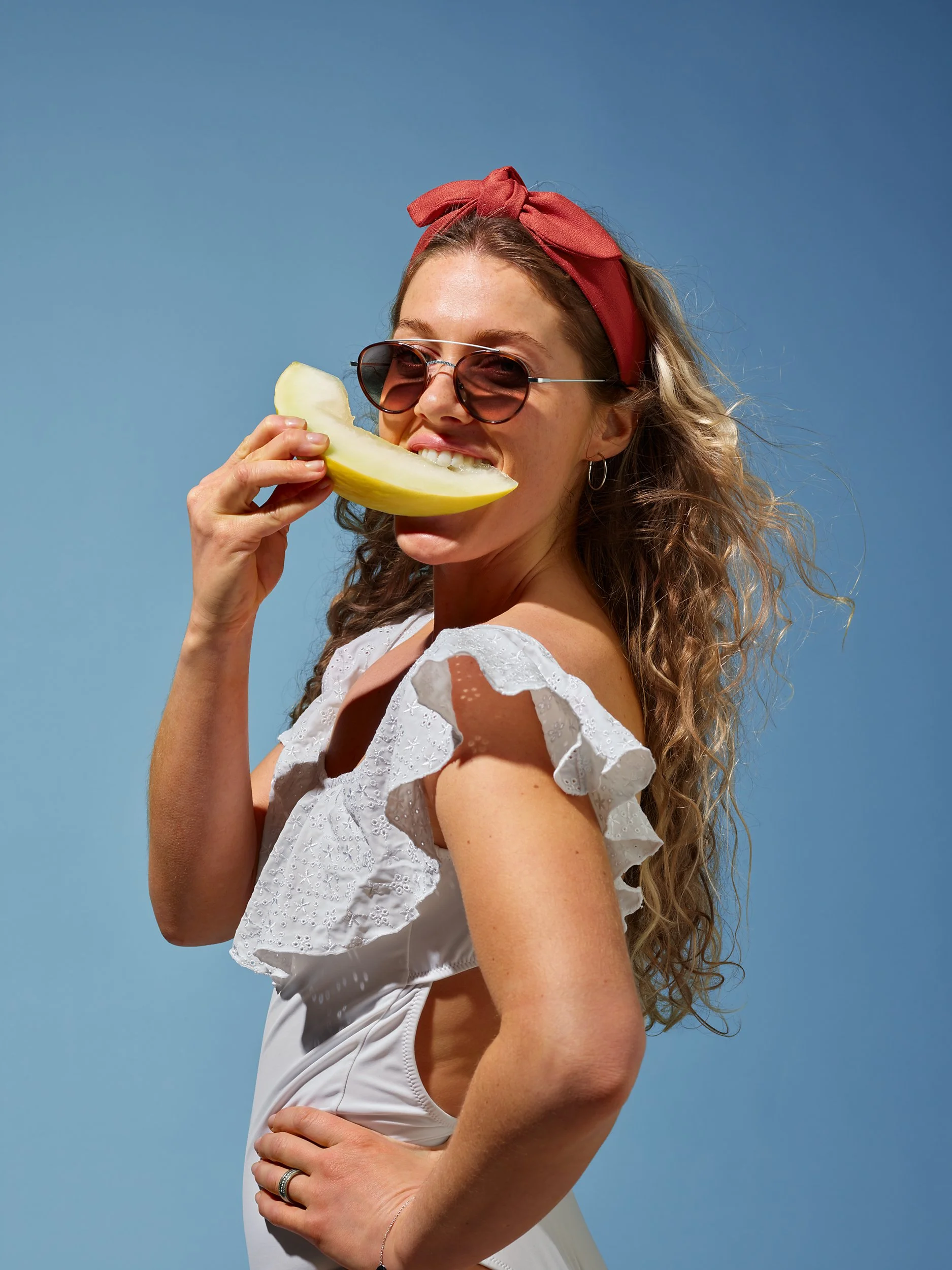A woman with curly hair, wearing sunglasses, a red headband, and a white dress, is smiling and holding a slice of melon near her mouth against a clear blue sky.