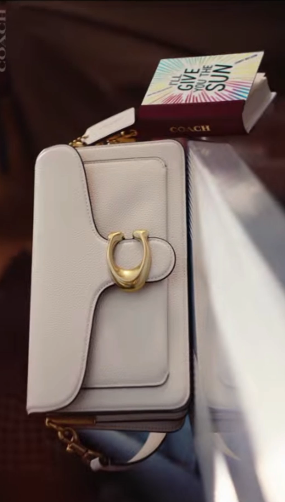 A white Coach handbag with a gold clasp, with a colorful box that says "The Give You the Sun" on top of it, placed on a dark surface.