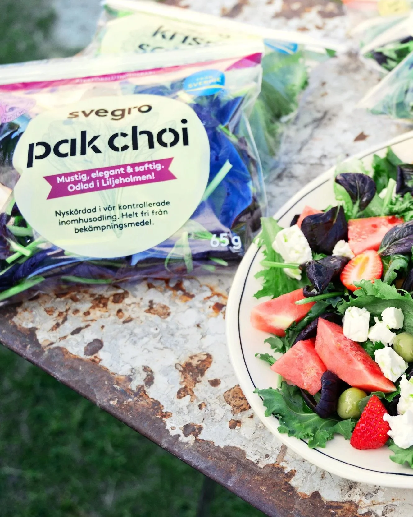 A large bowl of mixed fruit and salad greens, including strawberries, watermelon, grapes, feta cheese, and grilled pineapple slices, placed on a rustic outdoor table, with bags of pre-washed salad greens in the background.