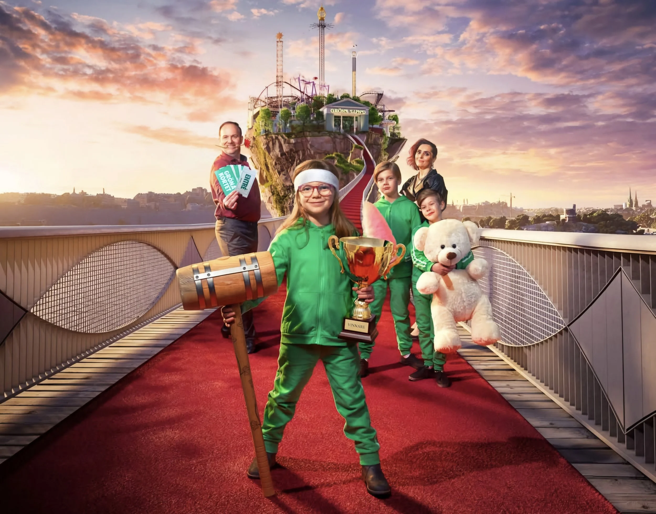 A group of people, including children and a woman, standing on a red carpet with a pirate-themed backdrop, holding trophies and plush toys, with an amusement park ride in the background at sunset.