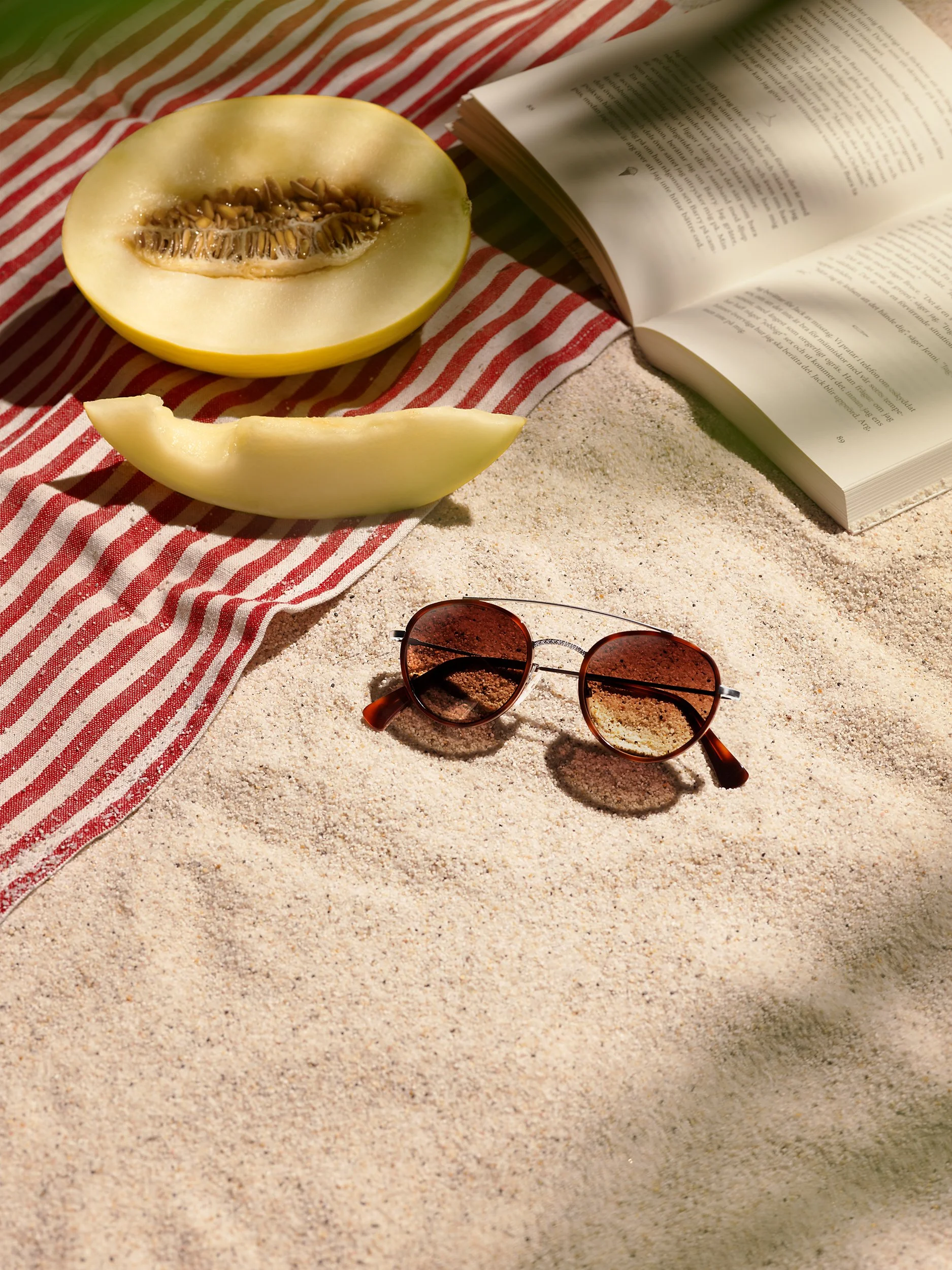 A half cantaloupe melon and a piece on a sandy surface, with an open book, a pair of sunglasses, and a red and white striped cloth.