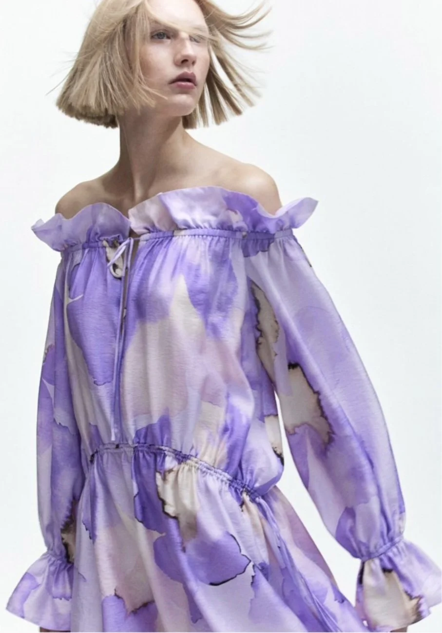 A woman with short blonde hair wearing a purple and white floral off-the-shoulder dress with ruffled sleeves.