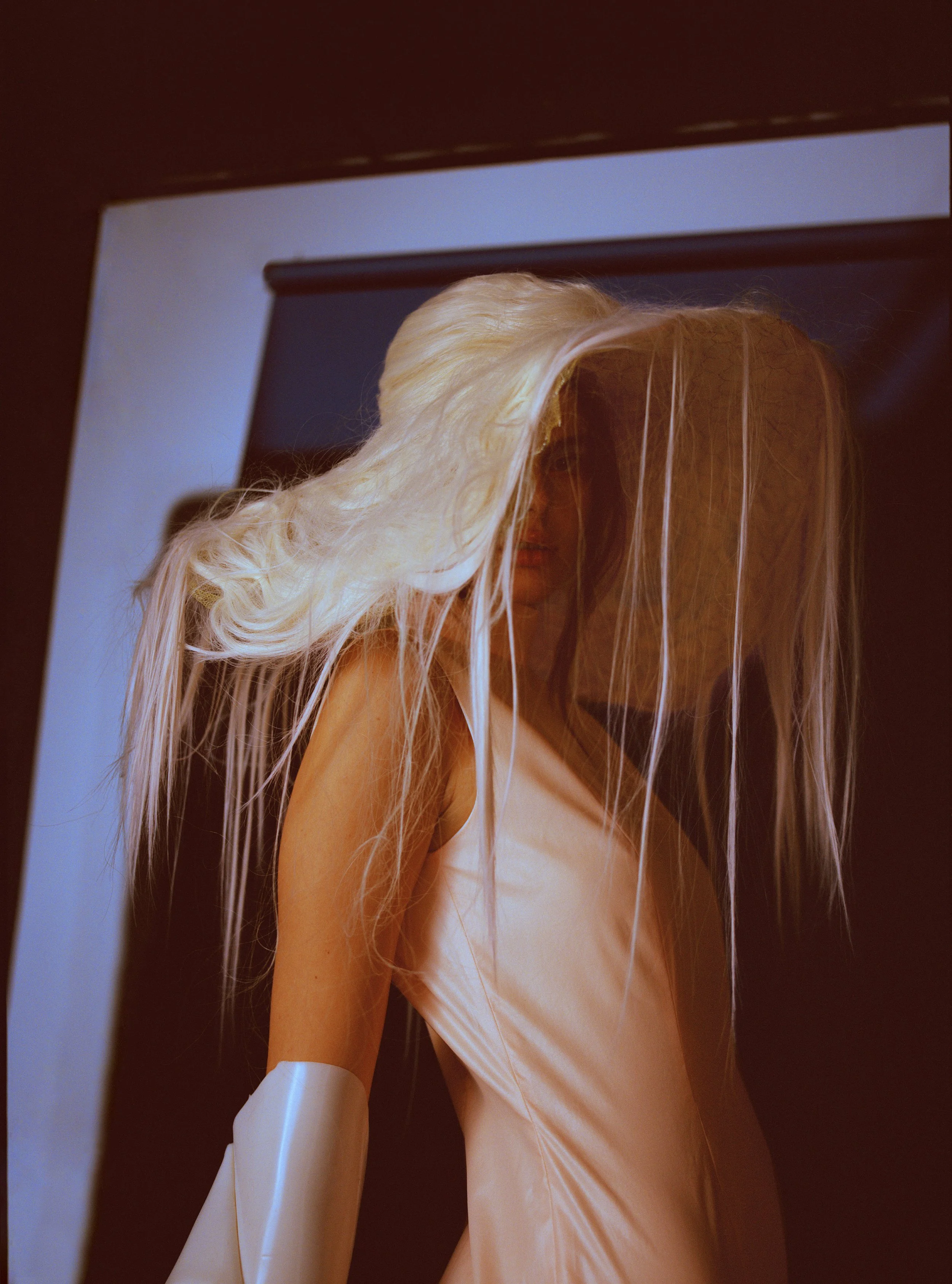 A person with long blonde hair wearing a beige dress and white gloves, standing in front of a dark background.