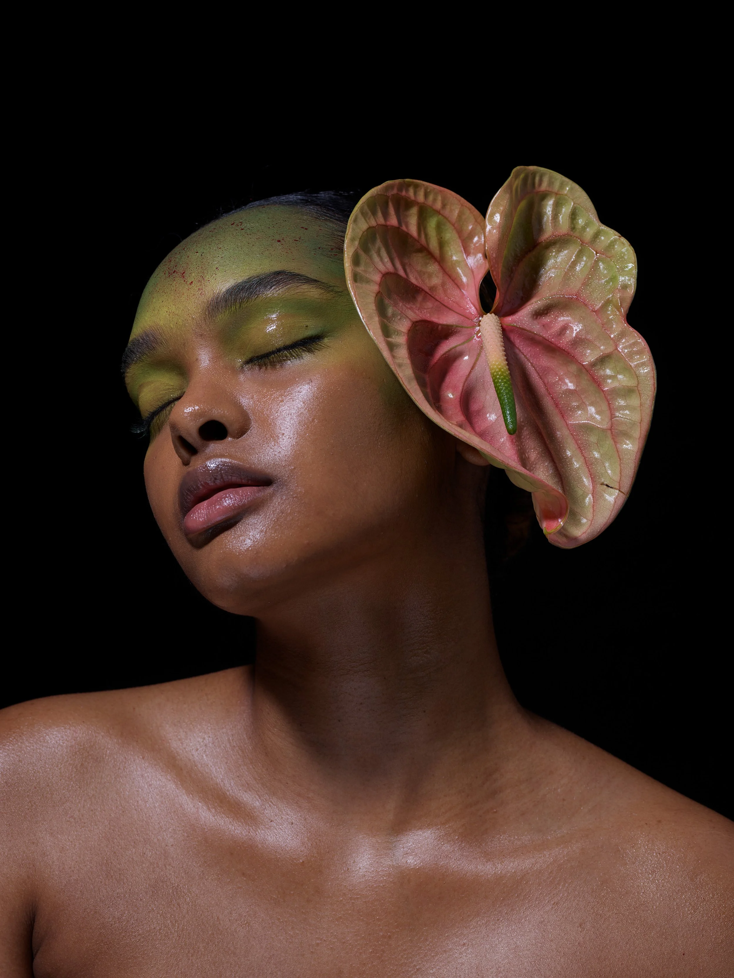 Close-up of a person with makeup and a large anthurium flower positioned near their face, dark background.