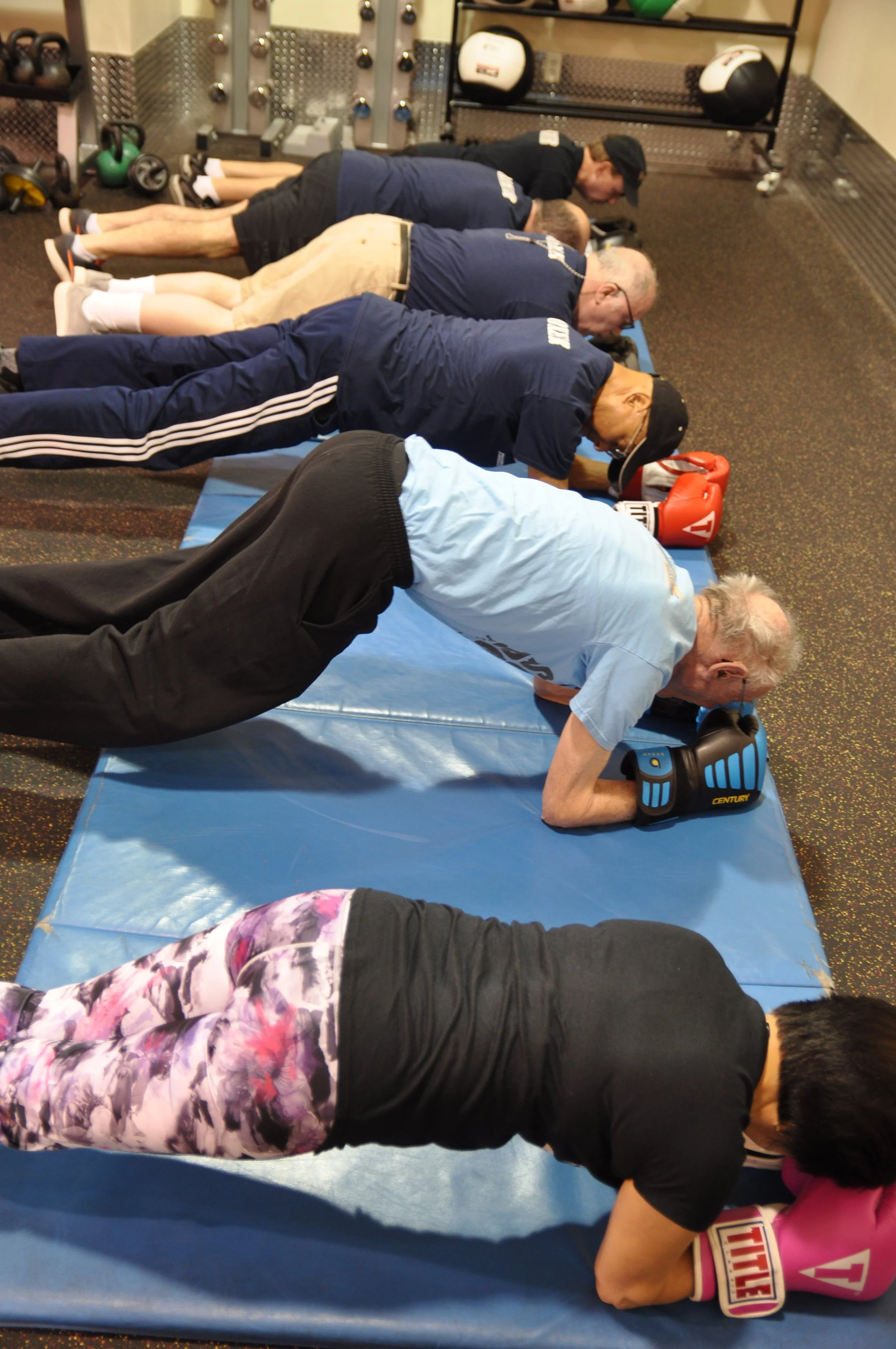 Group of people exercising in a gym, doing planks on mats, wearing boxing gloves.