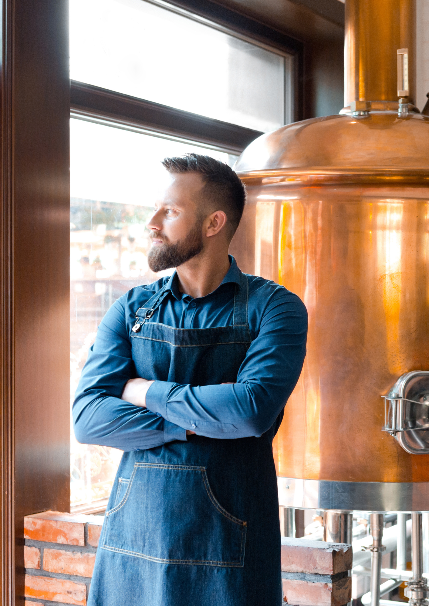 A man with a beard in a blue shirt and denim apron standing with arms crossed, looking out the window in a brewery.