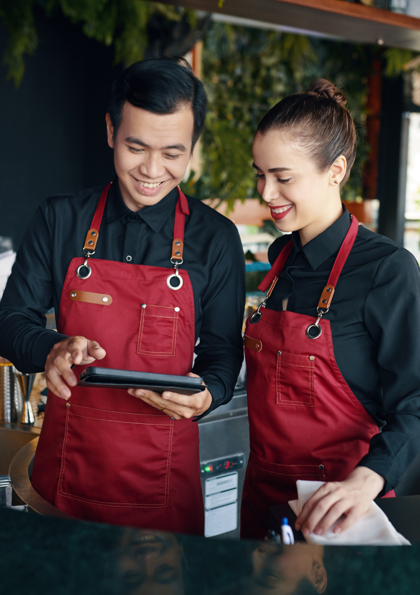 Two restaurant employees in black shirts and red aprons looking at a digital device, smiling, in a restaurant setting.