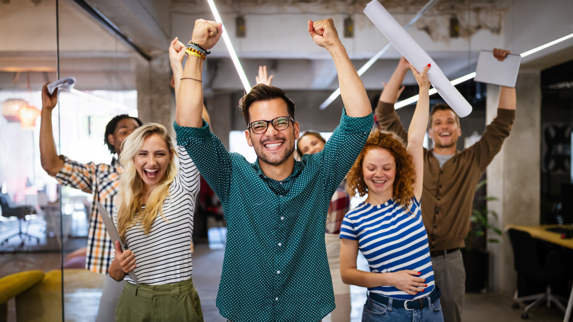 Group of five joyful people celebrating with raised arms in a modern office environment.