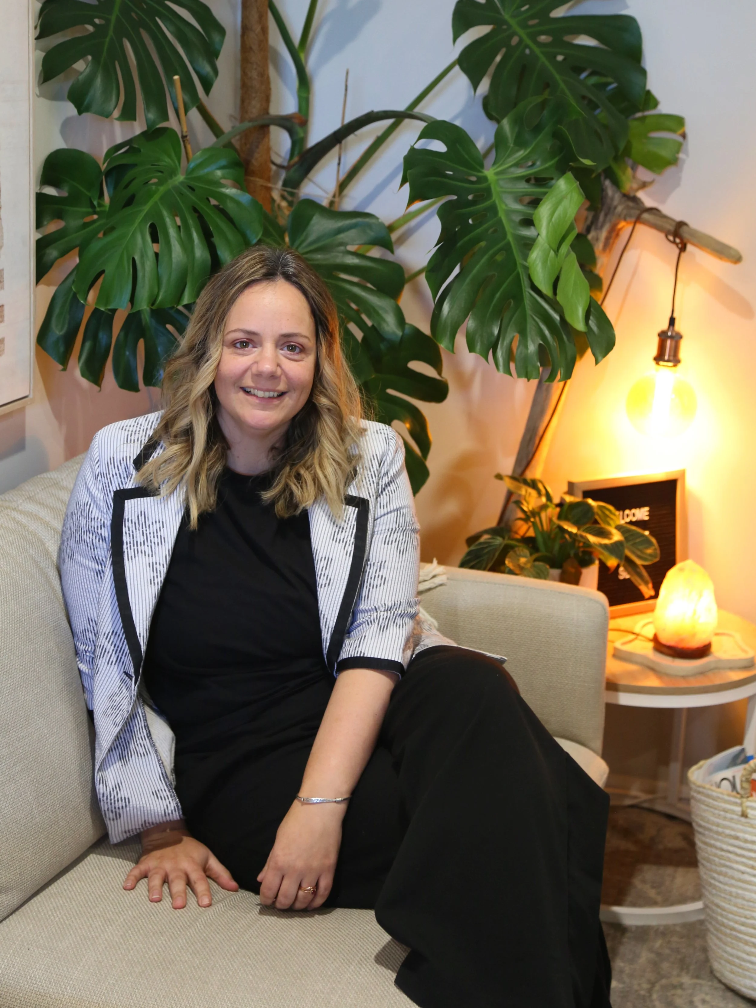 A woman with shoulder-length wavy blonde hair, wearing a black dress and a light-colored blazer with black trim, sitting on a beige sofa in a cozy, well-decorated room with large green monstera plants, a side table with a salt lamp, and a glowing light bulb hanging from the ceiling.