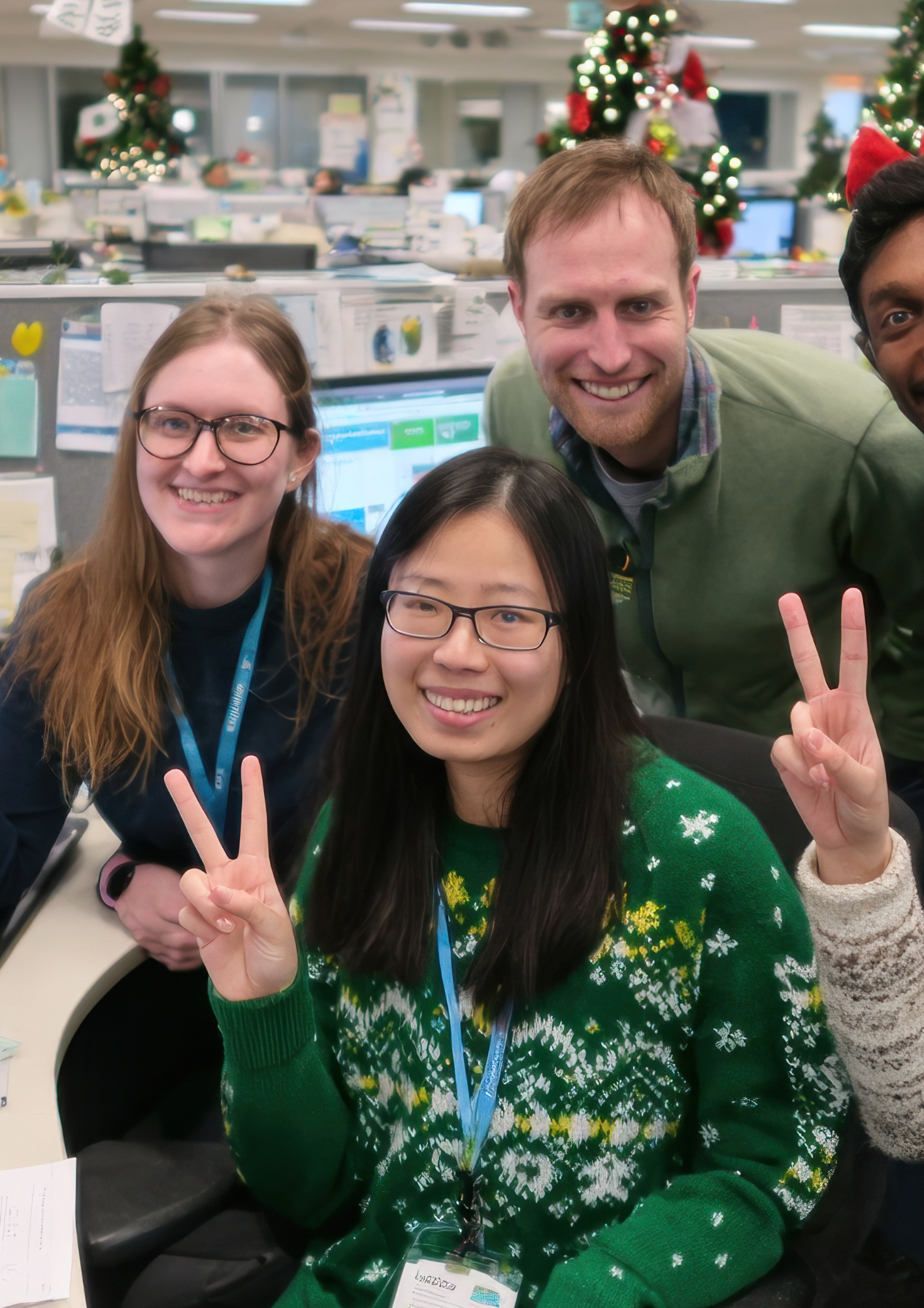 Group of five coworkers smiling and making peace signs in an office decorated with Christmas trees.
