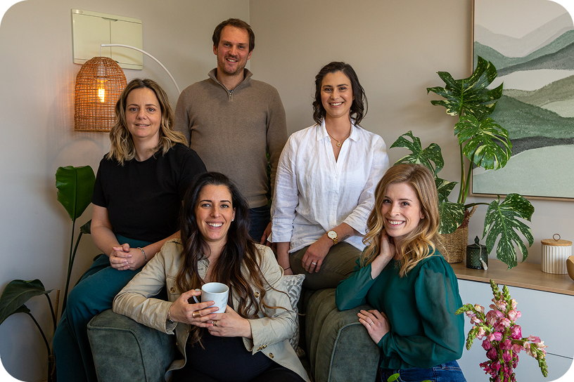 A group of five people, three women and two men, smiling and posing in a living room with plants and modern decor.