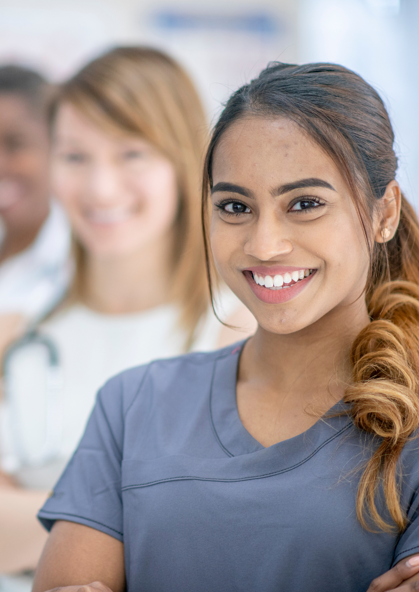 Smiling young female healthcare professional in scrubs with other medical staff in background.