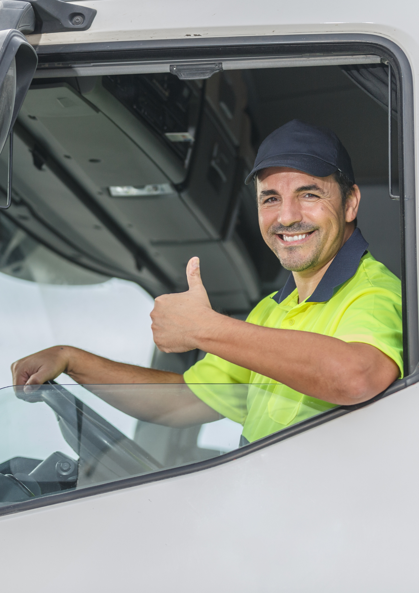 A smiling man wearing a high visibility safety shirt and cap, sitting in the driver's seat of a truck, giving a thumbs up.