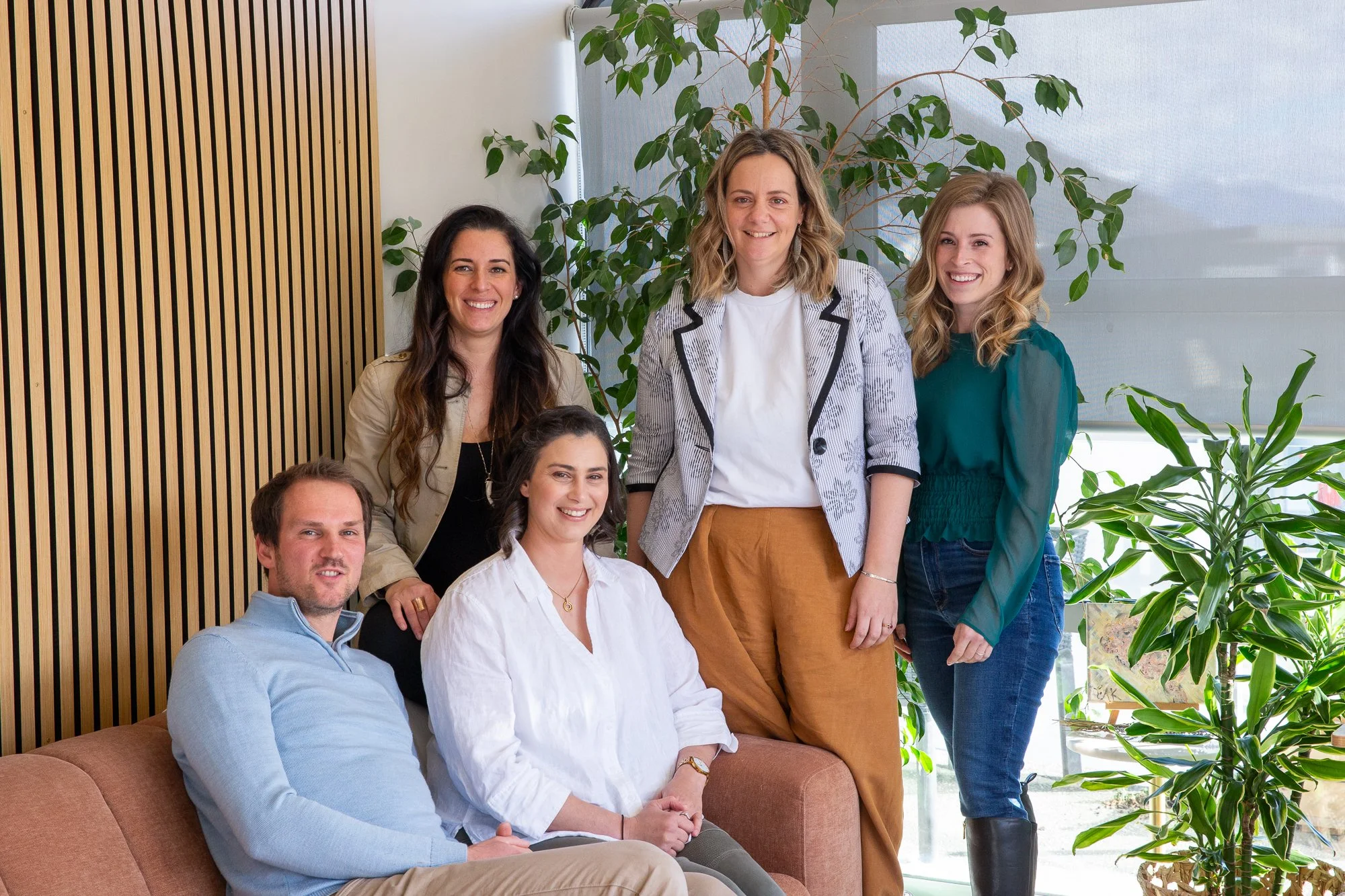Six people, five women and one man, smiling and posing together in an indoor setting with plants and wooden wall paneling.