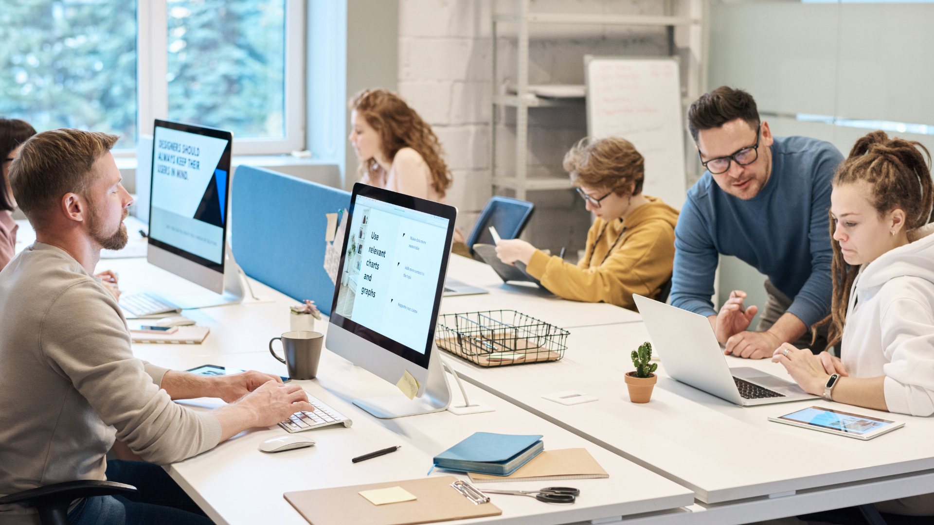 Office meeting room with six people working on computers and tablets at a long table, with a large window in the background.