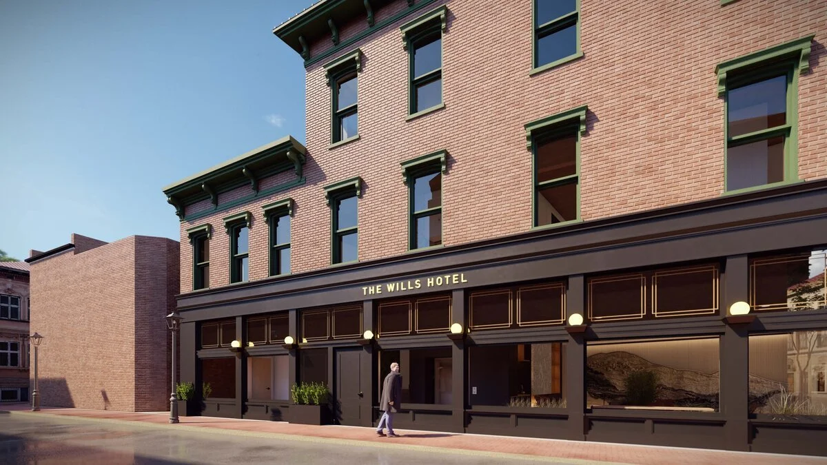 Rendering of The Wills Hotel exterior featuring a brick building facade with green window trims, large windows on the ground floor, and a person walking on the sidewalk in front, under a clear sky.