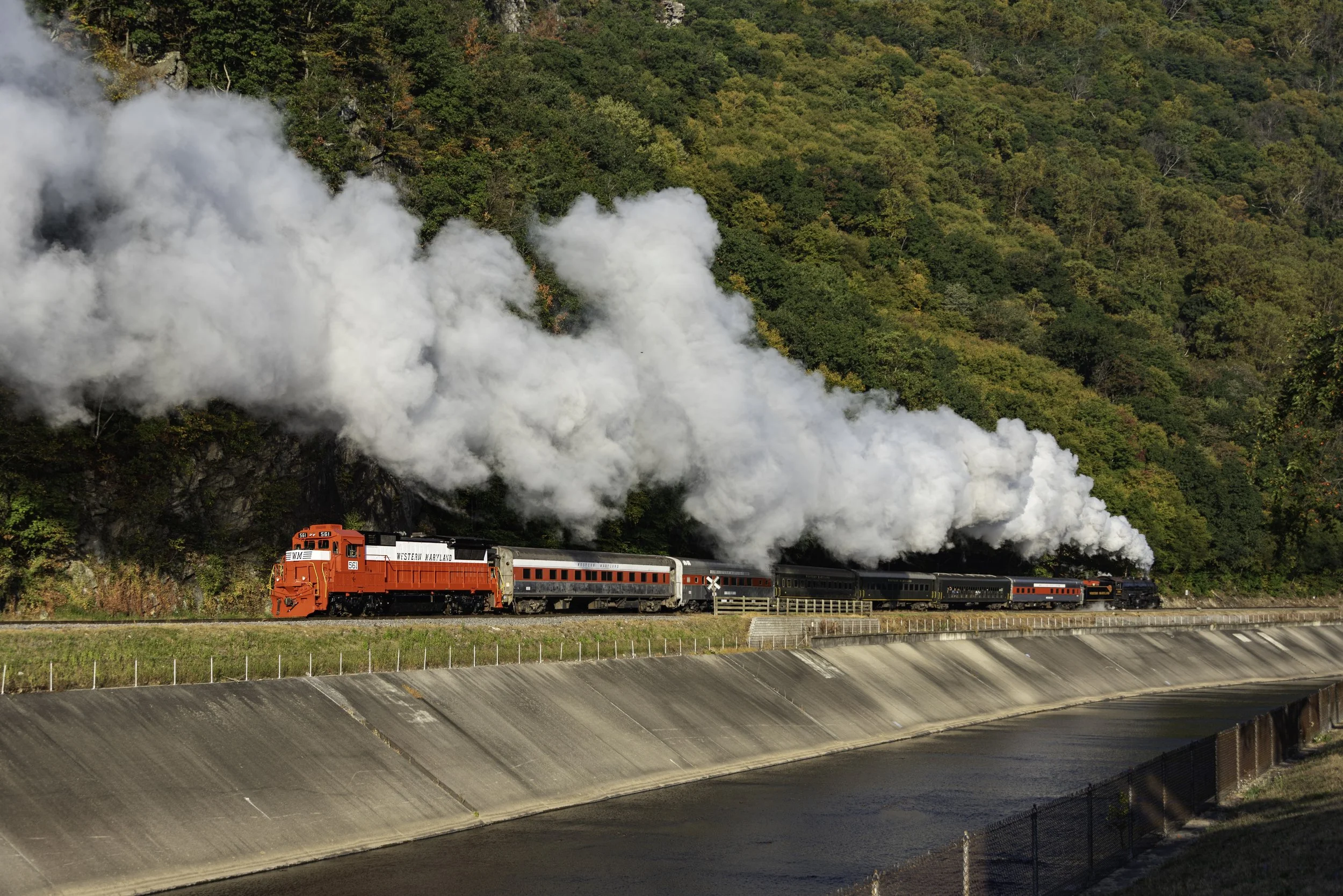 A red and black train moving along tracks next to a large concrete dam, with a backdrop of green forested hills and white smoke or steam coming from the train.