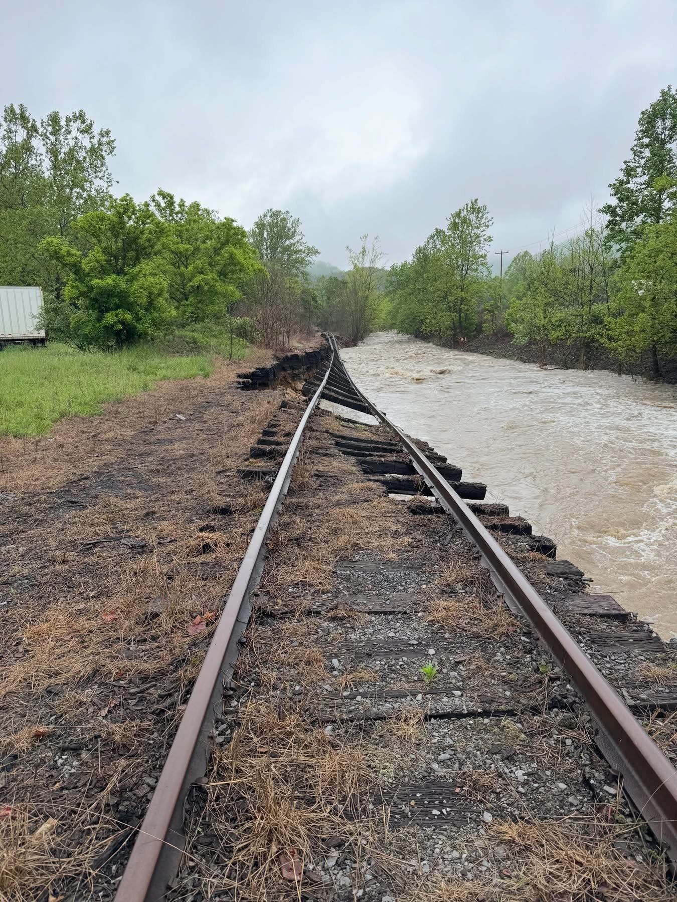 Flooded railway tracks partially washed away by floodwaters, with debris and damaged ties, surrounded by green trees and cloudy sky.