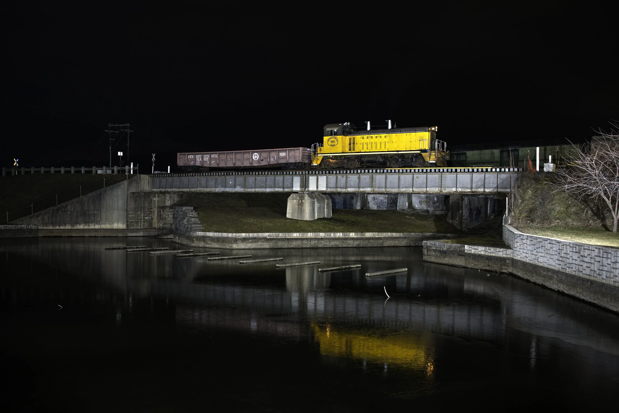 Nighttime photo of a yellow train engine with a purple freight car and green railcars on a bridge over a river. The scene is dark with reflections in the water below.