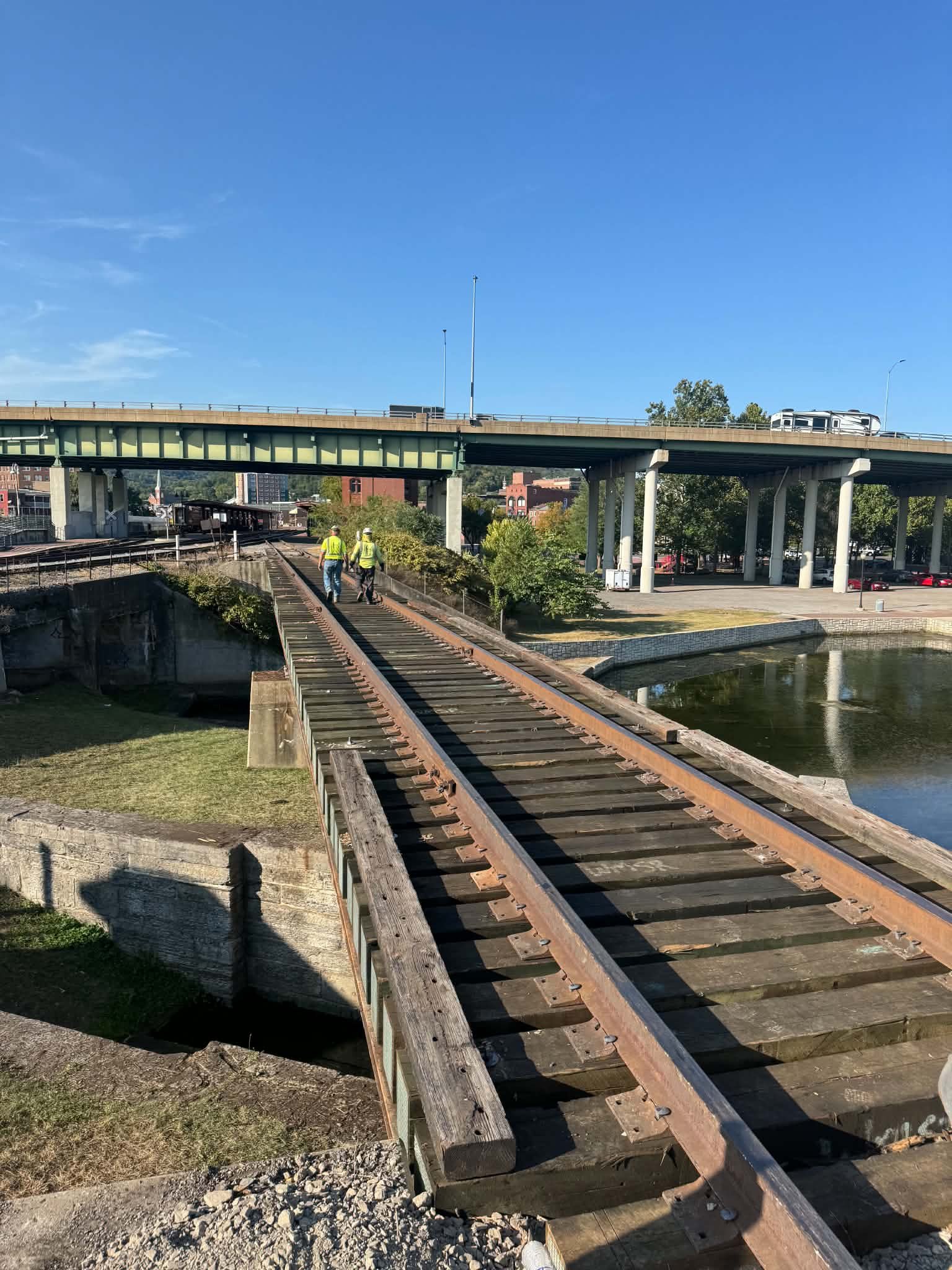 Three workers wearing safety vests and helmets walking on a damaged railway track underneath a bridge over a river on a sunny day.