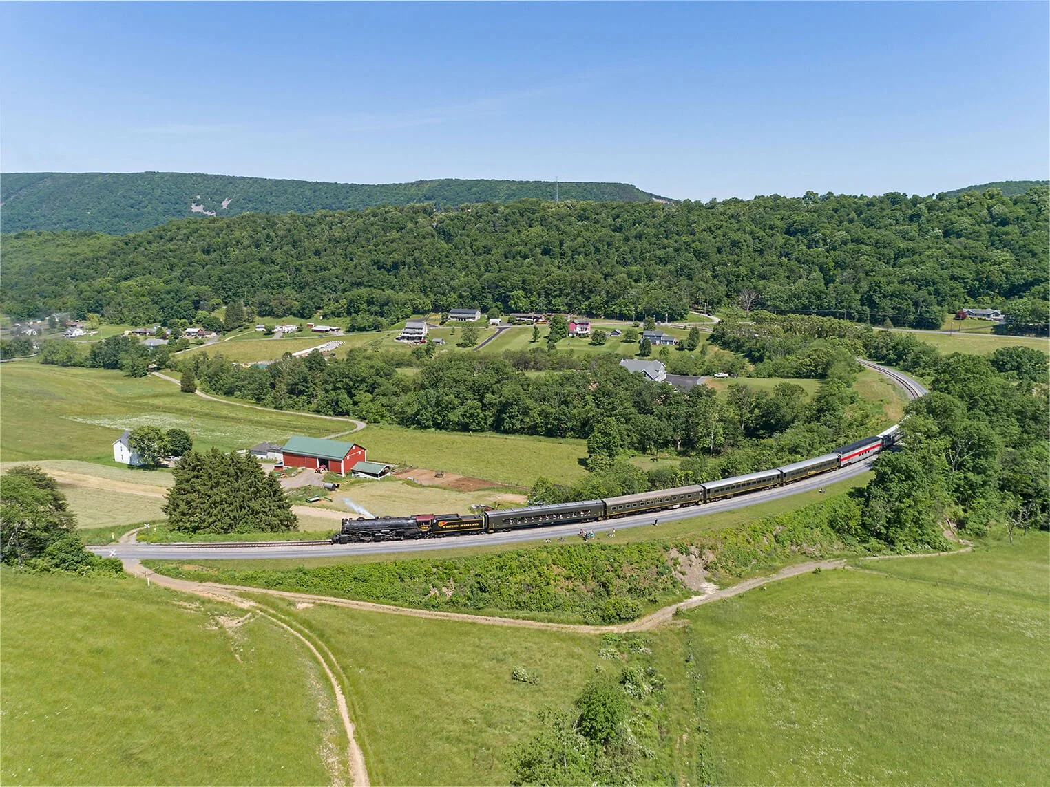A steam train traveling through lush green countryside with houses and trees, under a blue sky with distant hills.