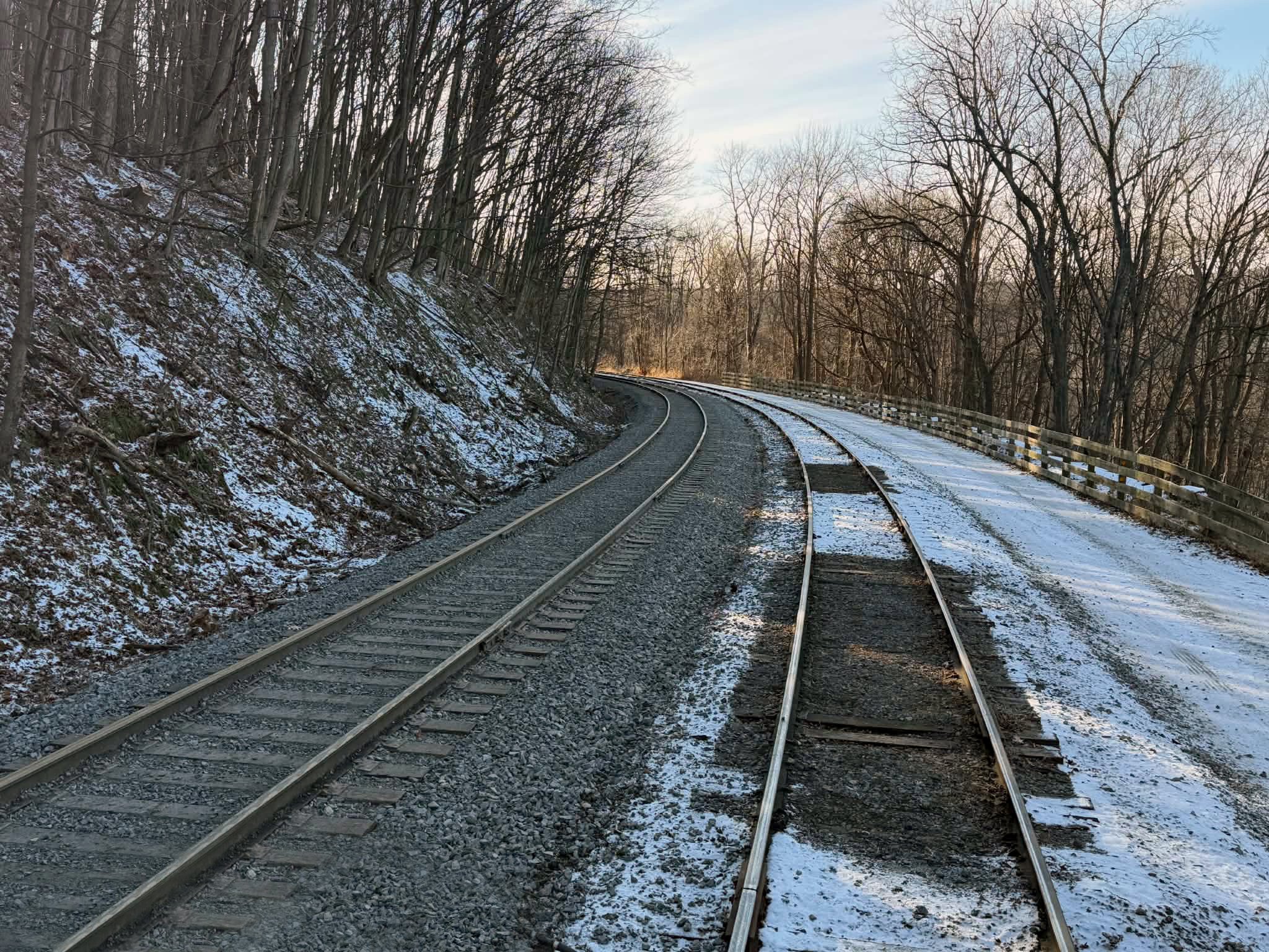 Curving train tracks in a winter landscape with snow on the ground and leafless trees on both sides, a fence along the right side, and a partly cloudy sky.