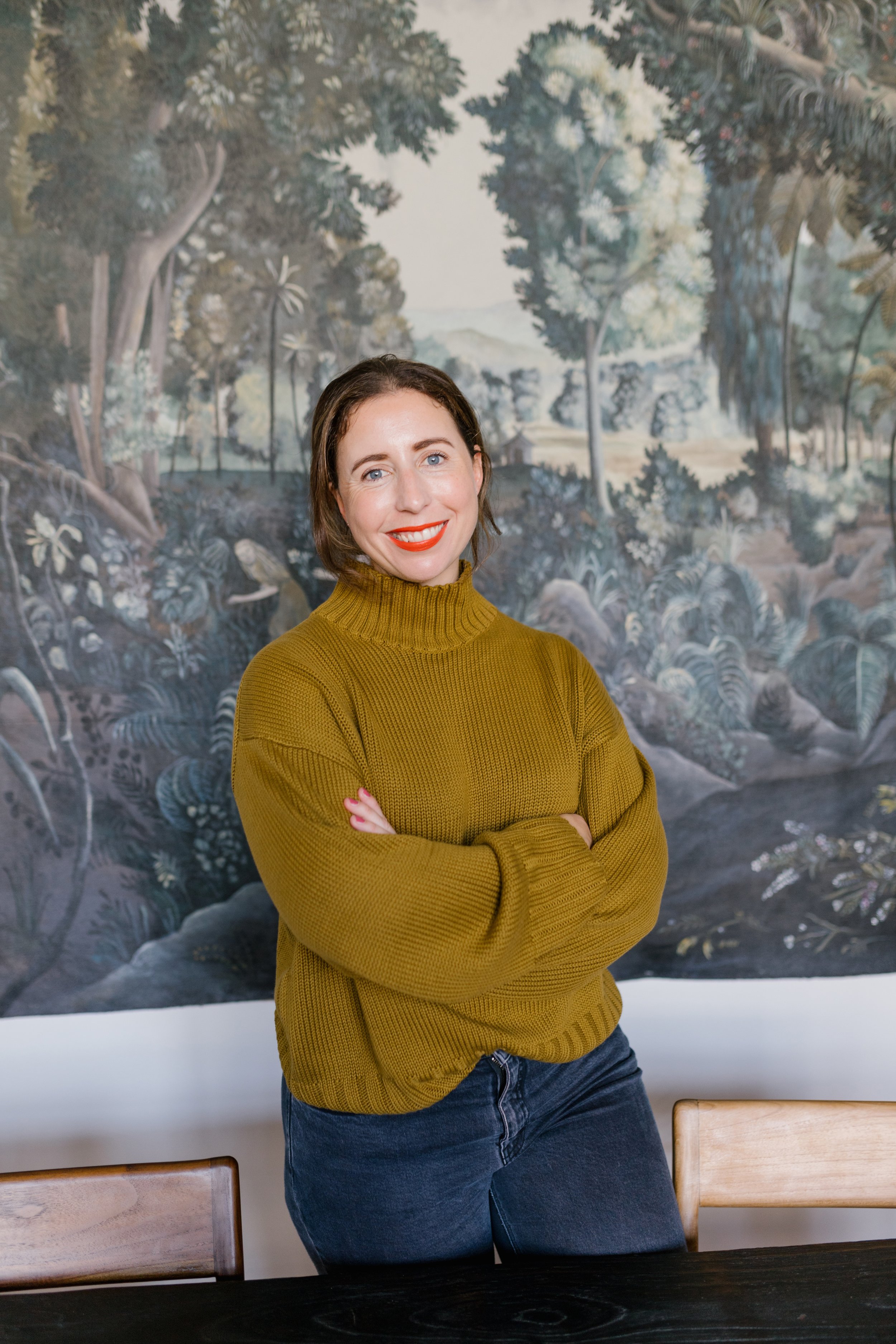 A woman in a mustard yellow sweater and dark jeans standing with crossed arms, smiling in front of a large landscape painting of a forest scene with trees and mountains.
