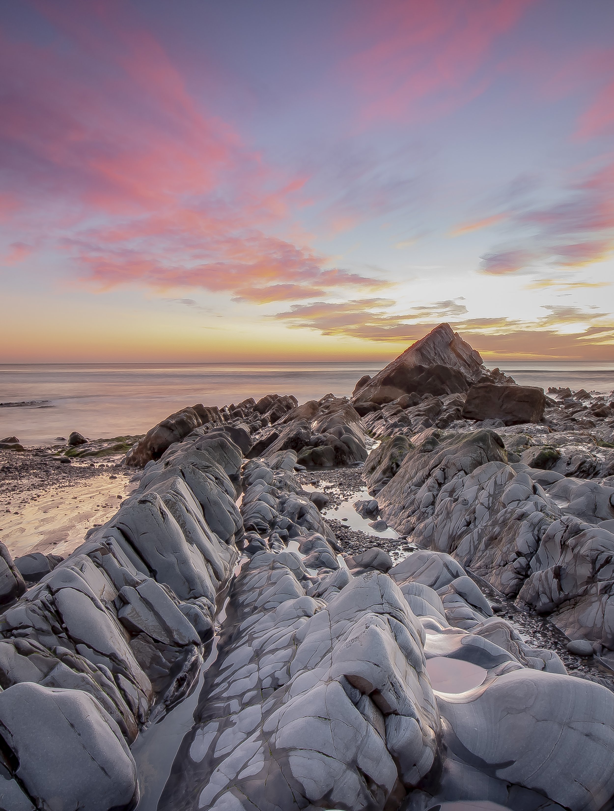 Photo by Kim Stone, North Devon coast, A scenic view of a rocky shoreline at sunset or sunrise with pink and purple clouds in the sky and calm water in the background.