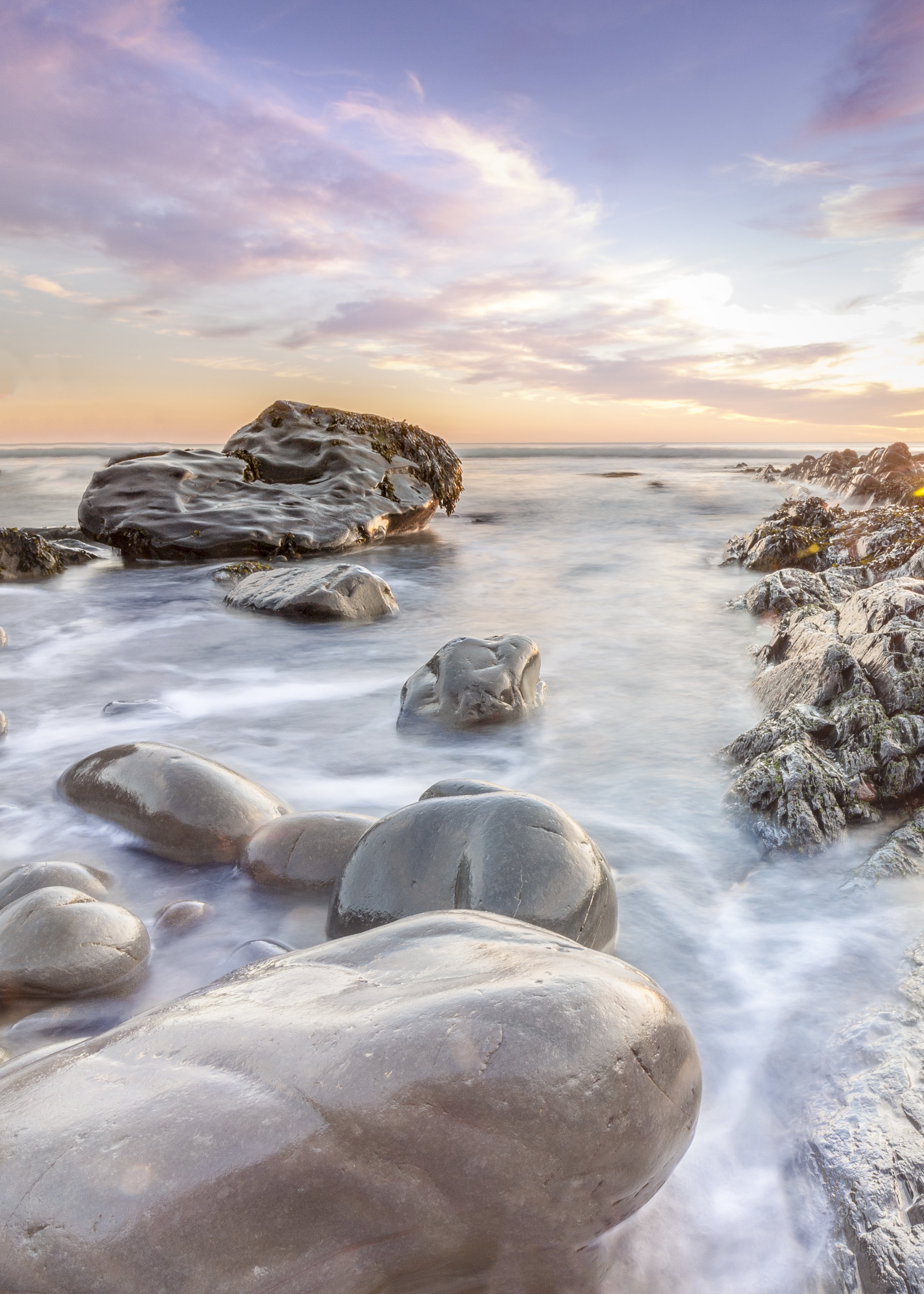 Photo by Kim Stone of Sunset over Westward Ho! rocky shoreline with large, wet stones and calm ocean waves.