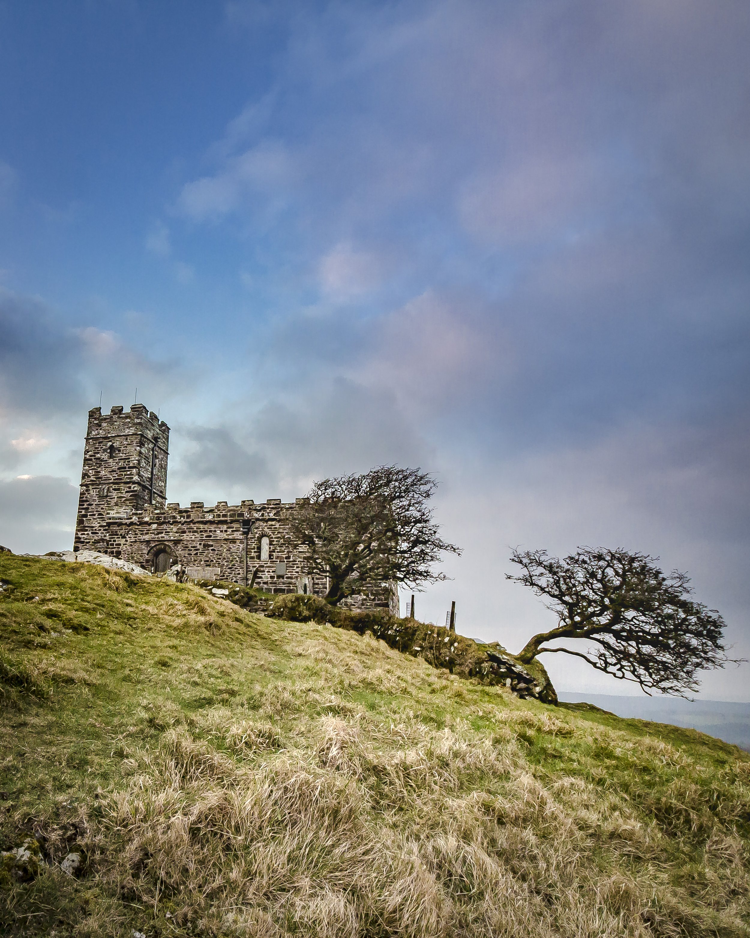 Photo by Kim Stone of Brentor Church, Dartmoor on a grassy hill with two leafless trees, under a partly cloudy sky.