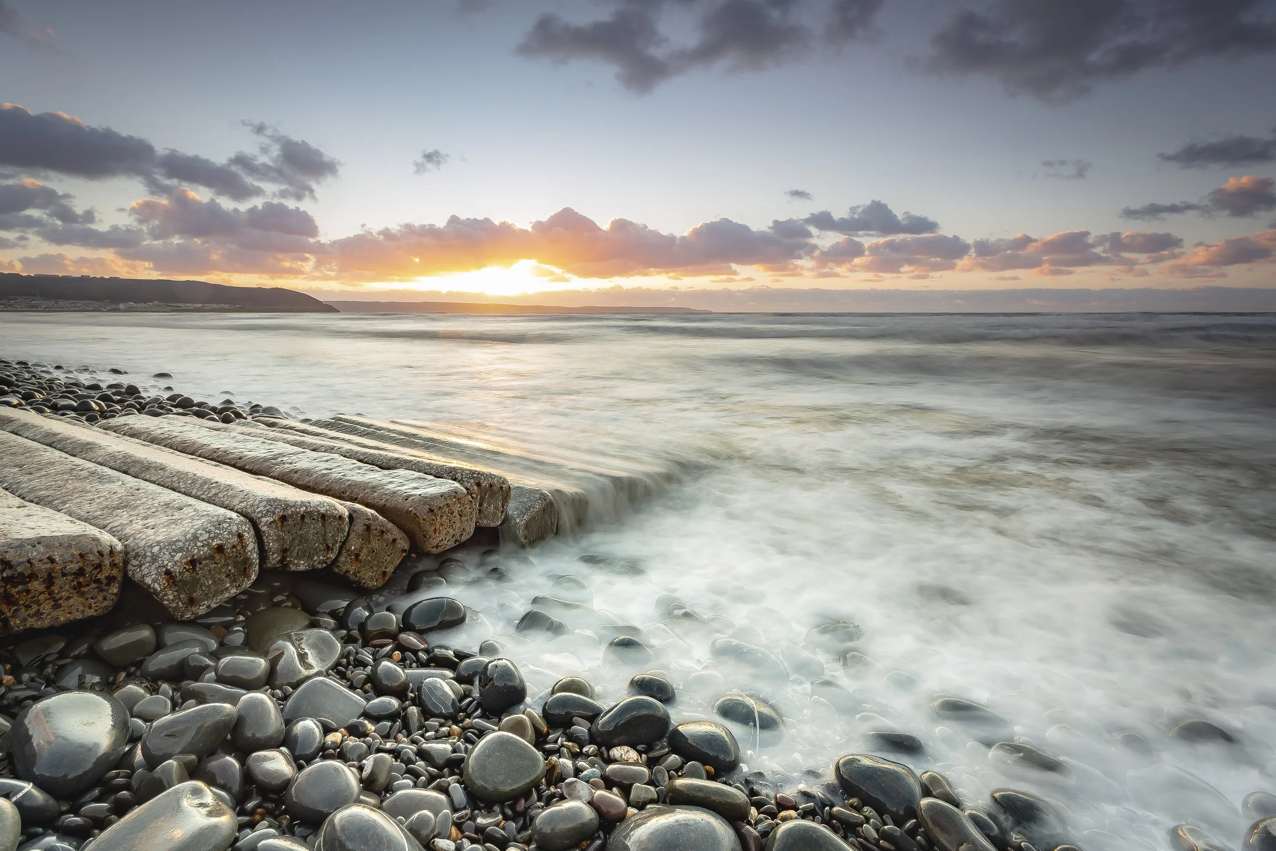 Photo by Kim Stone of Westward Ho! seascape at sunrise with a stone and concrete pier extending into the ocean, calm water, cloudy sky, and distant land on the horizon.