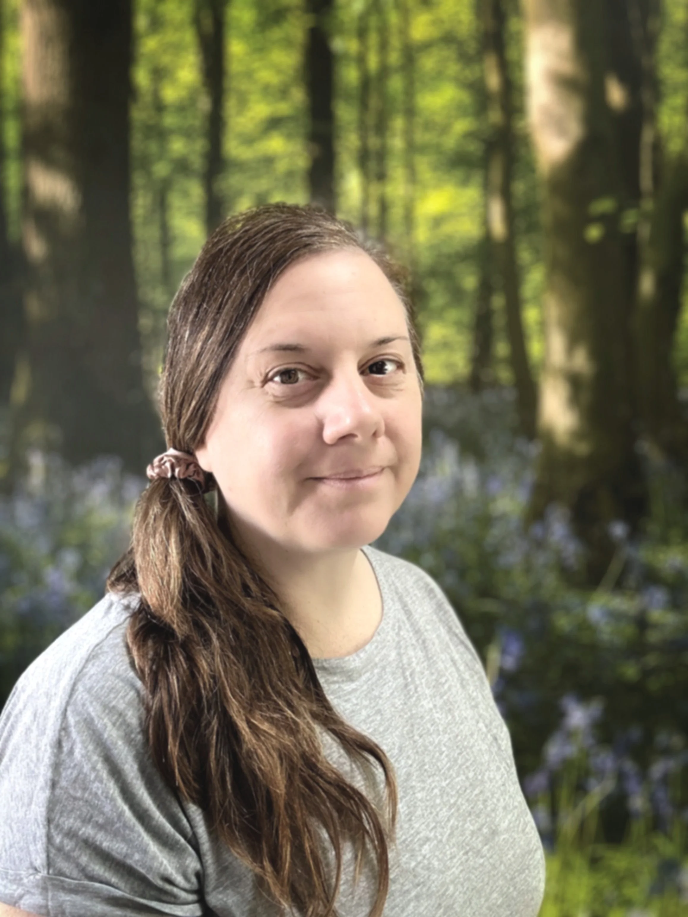 Kim Stone, Mindfulness Teacher with long brown hair tied in a ponytail, wearing a grey t-shirt, standing outdoors in a forest with trees and greenery in the background.