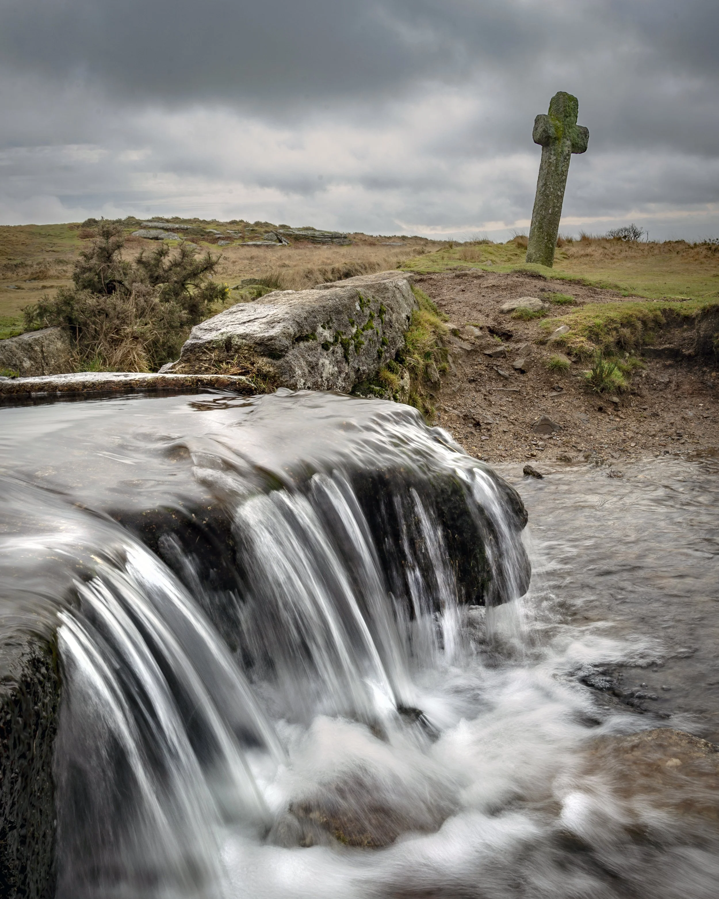 Photo by Kim Stone A small waterfall on Dartmoor flowing over rocks in a rural landscape with a stone cross in the background under cloudy skies.