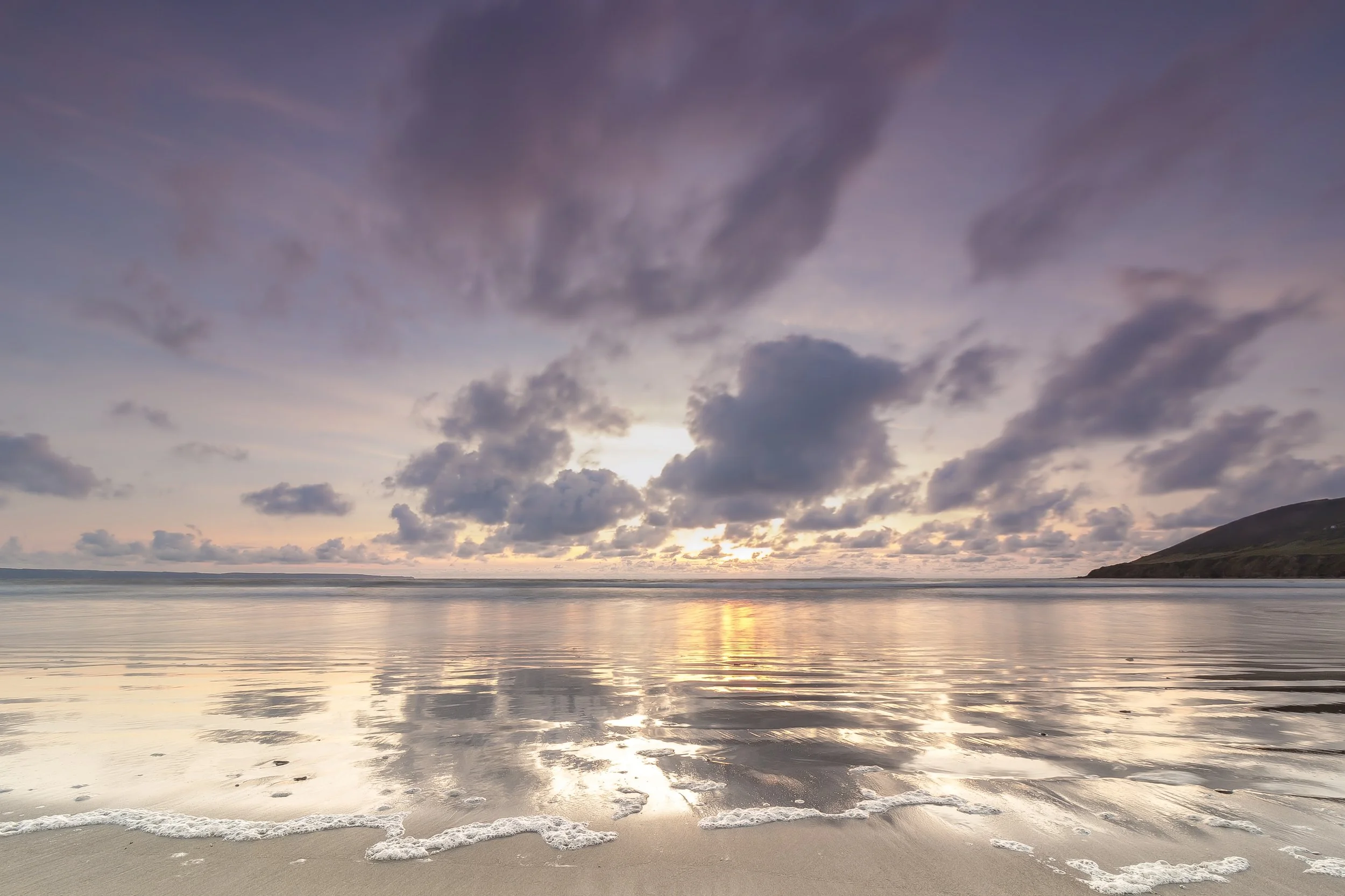 Photo by Kim Stone Sunset over a calm Saunton Sands beach with cloudy sky reflecting on the water and a distant hill on the right.