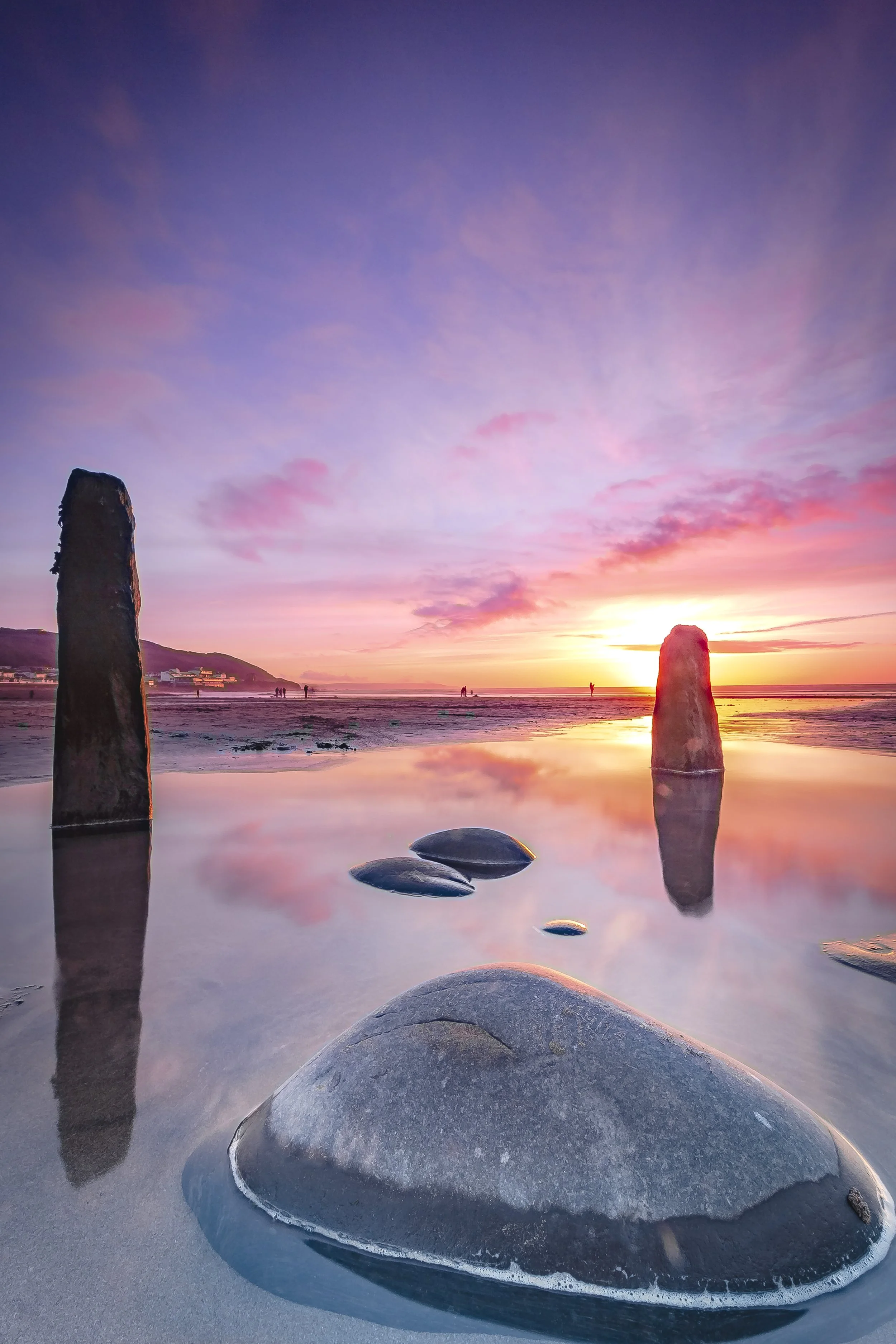 Photo by Kim Stone Westward Ho! Sunset over a calm beach with rocks and wooden posts in the shallow water, pink and purple sky with a few clouds, distant people and hills.